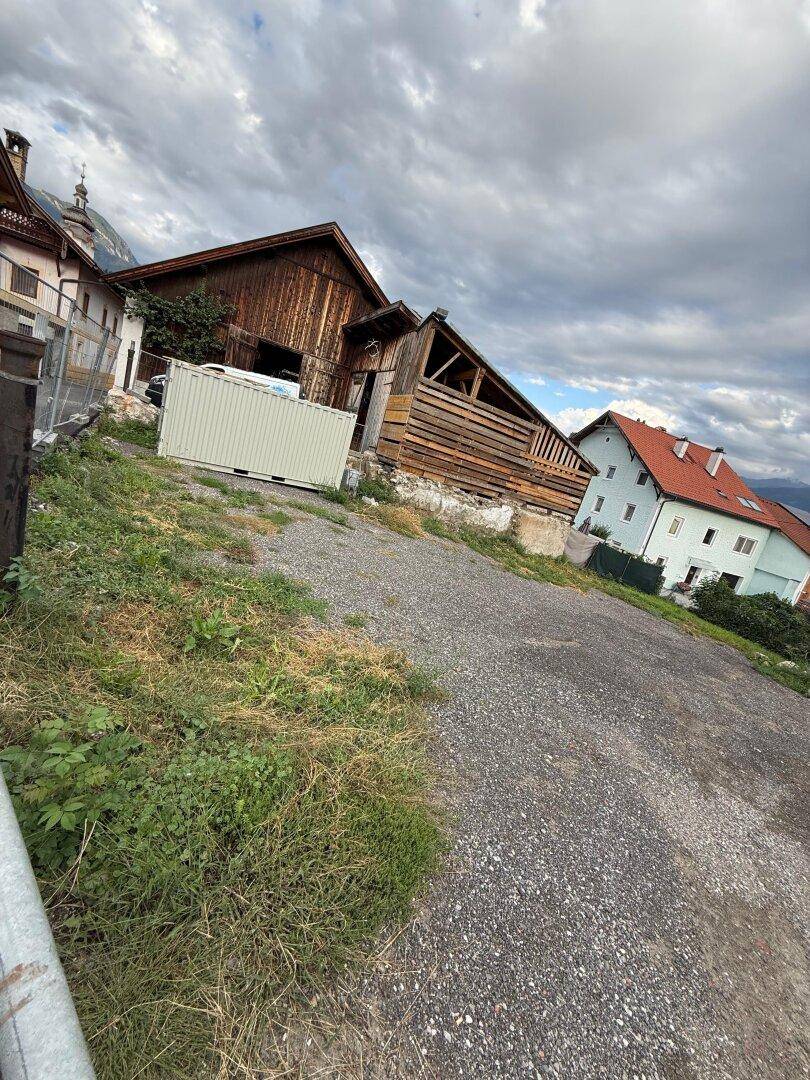 Schotterfläche eines Grundstücks mit Holzscheune und Container, umgeben von Häusern und Bergen.