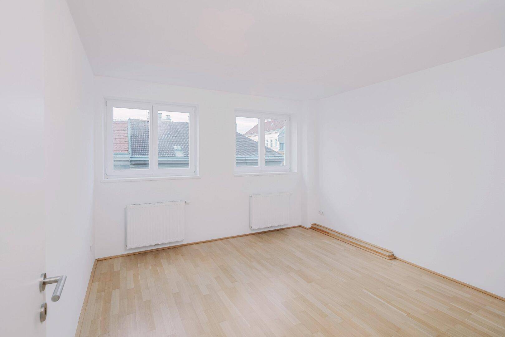 Helles Schlafzimmer mit Parkettboden, weißen Wänden und zwei Fenstern mit Blick auf Dächer.