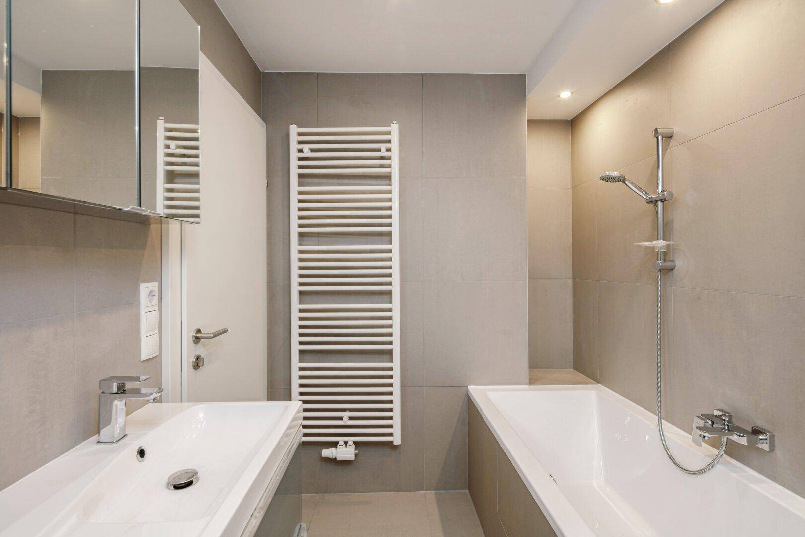 Modernes Badezimmer mit Badewanne, Waschbecken, beheiztem Handtuchhalter und grauen Fliesen.