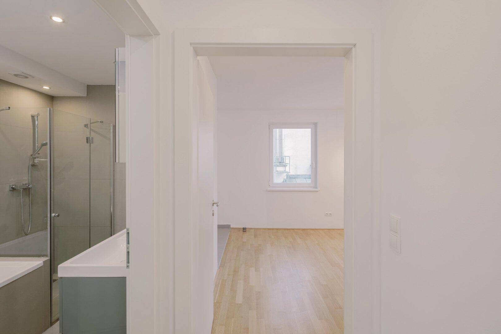 Blick aus einem Badezimmer in einen hellen Raum mit Parkettboden und Fenster.