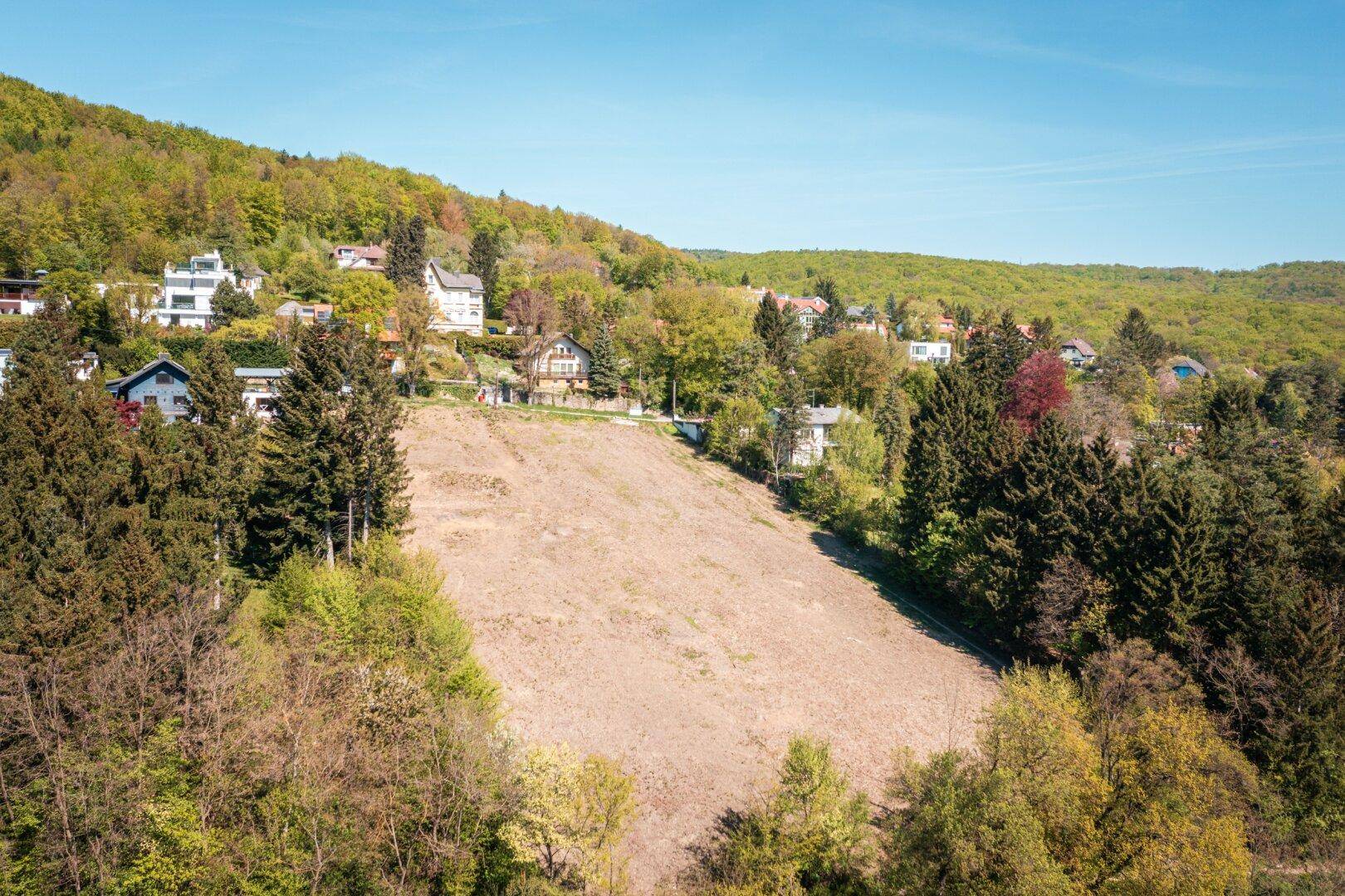 Luftaufnahme eines großen Baugrundstücks umgeben von bewaldeten Hügeln und Wohnhäusern.
