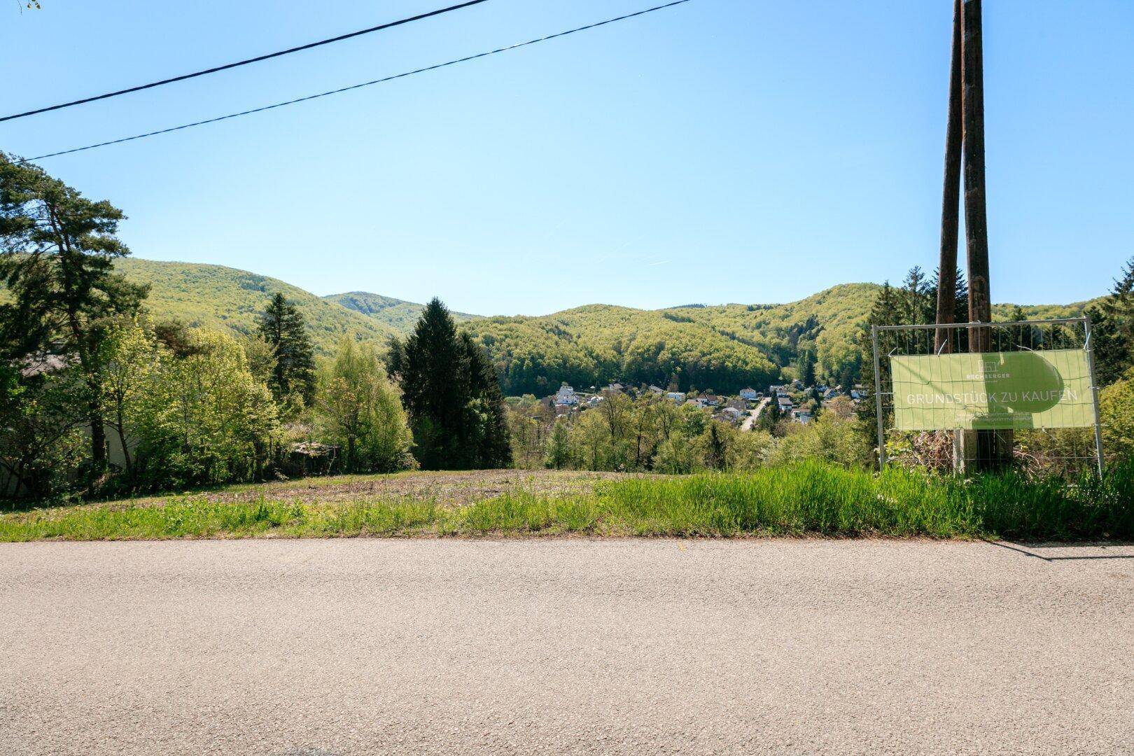 Blick von der Straße auf ein Baugrundstück mit einem Verkaufsschild und bewaldeten Hügeln im Hintergrund.