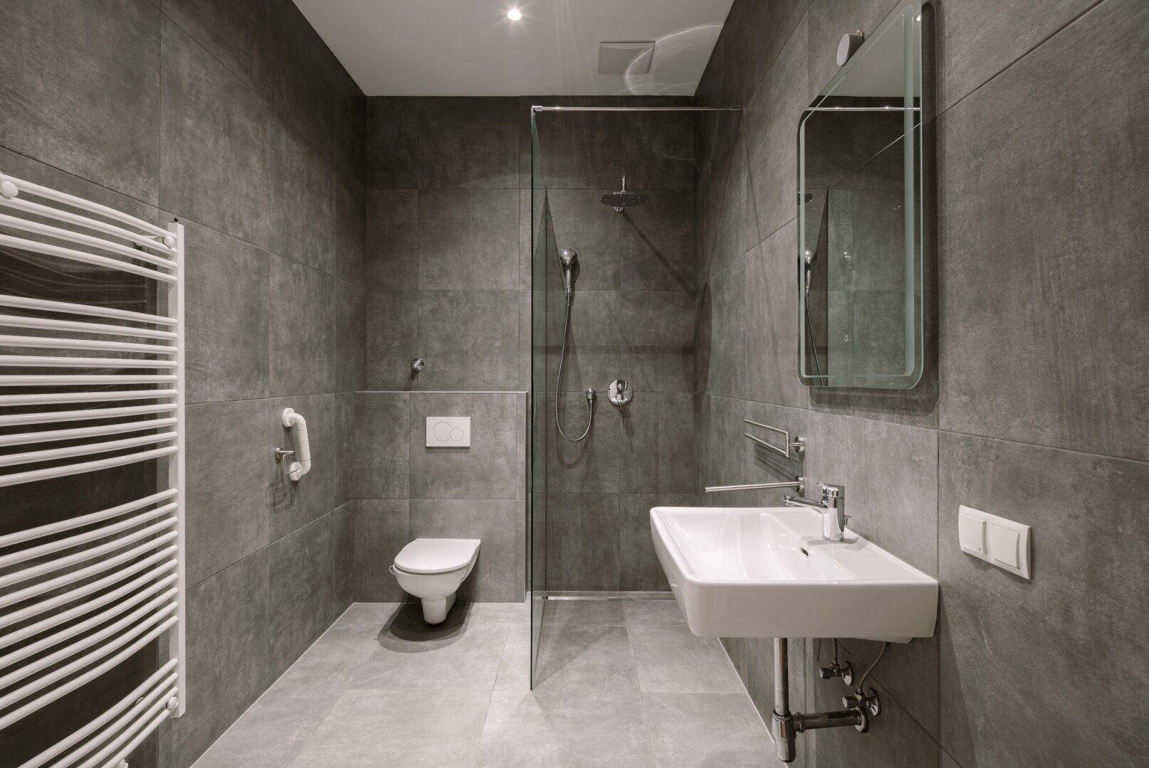 Modernes Badezimmer mit grauen Fliesen, ebenerdiger Dusche, Wand-WC und Haltegriffen.