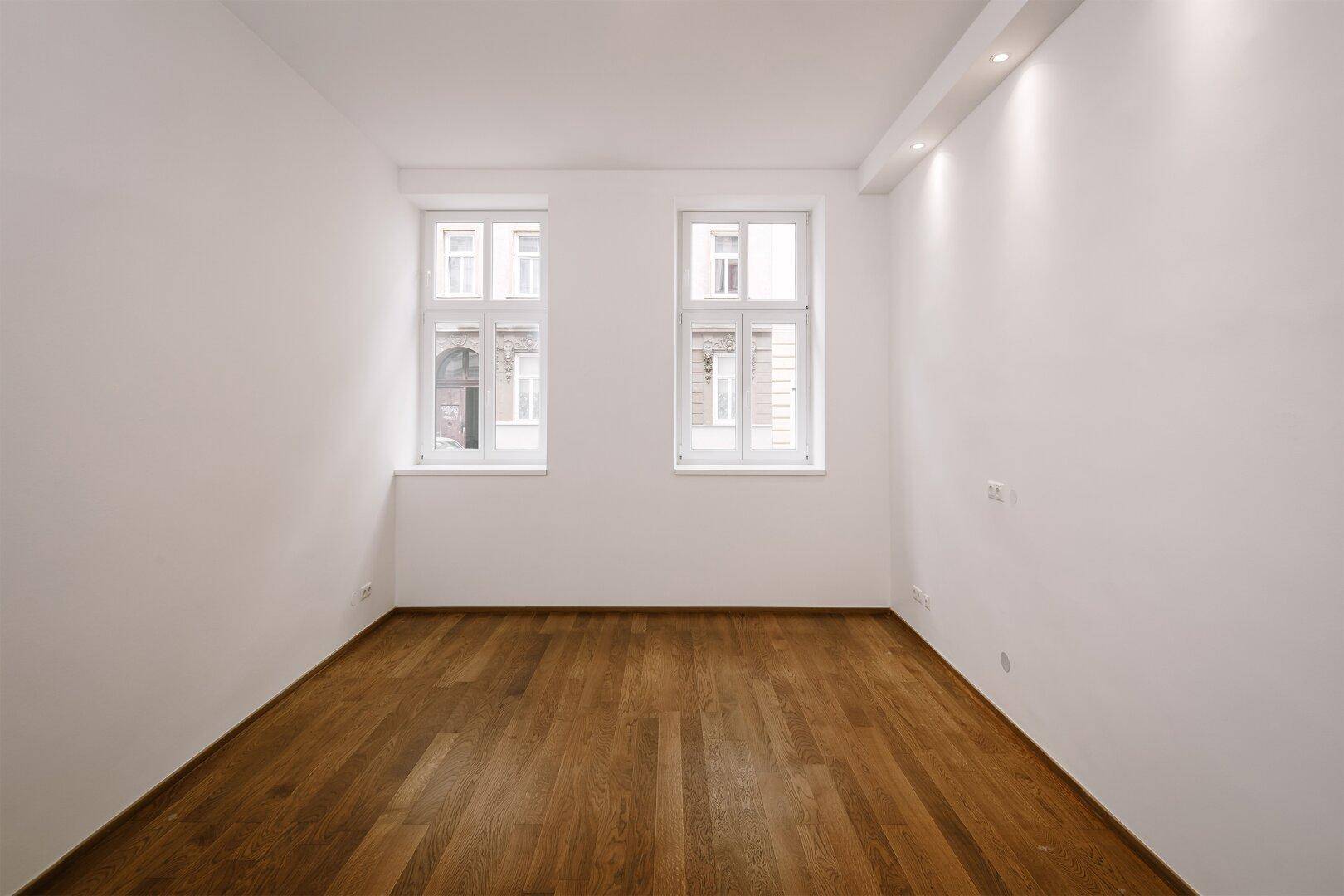 Geräumiger, heller Raum mit zwei großen Fenstern und elegantem Holzboden.