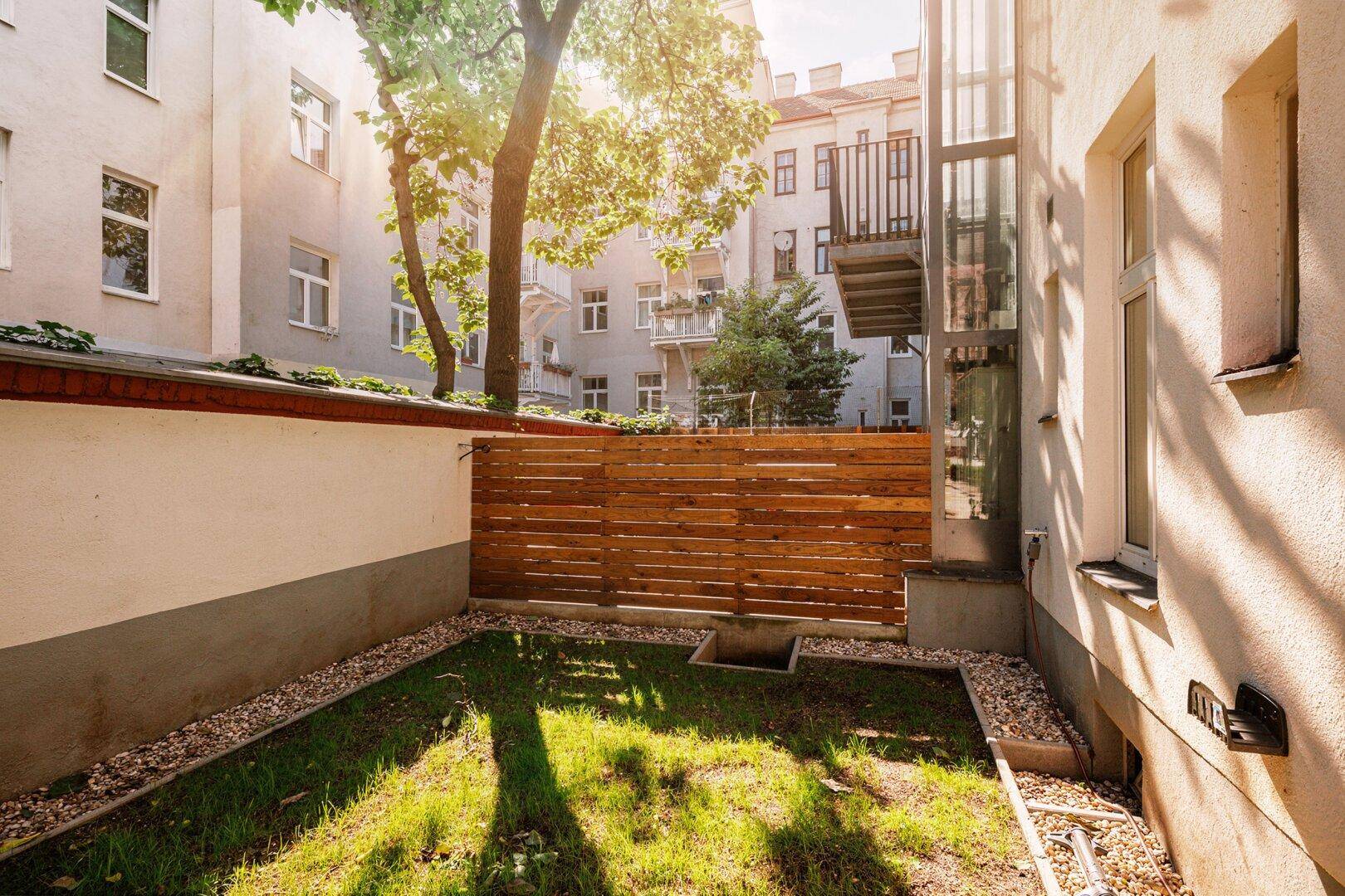 Sonniger Gartenbereich mit Rasenfläche, Holzabtrennung und Blick auf den Aufzug.