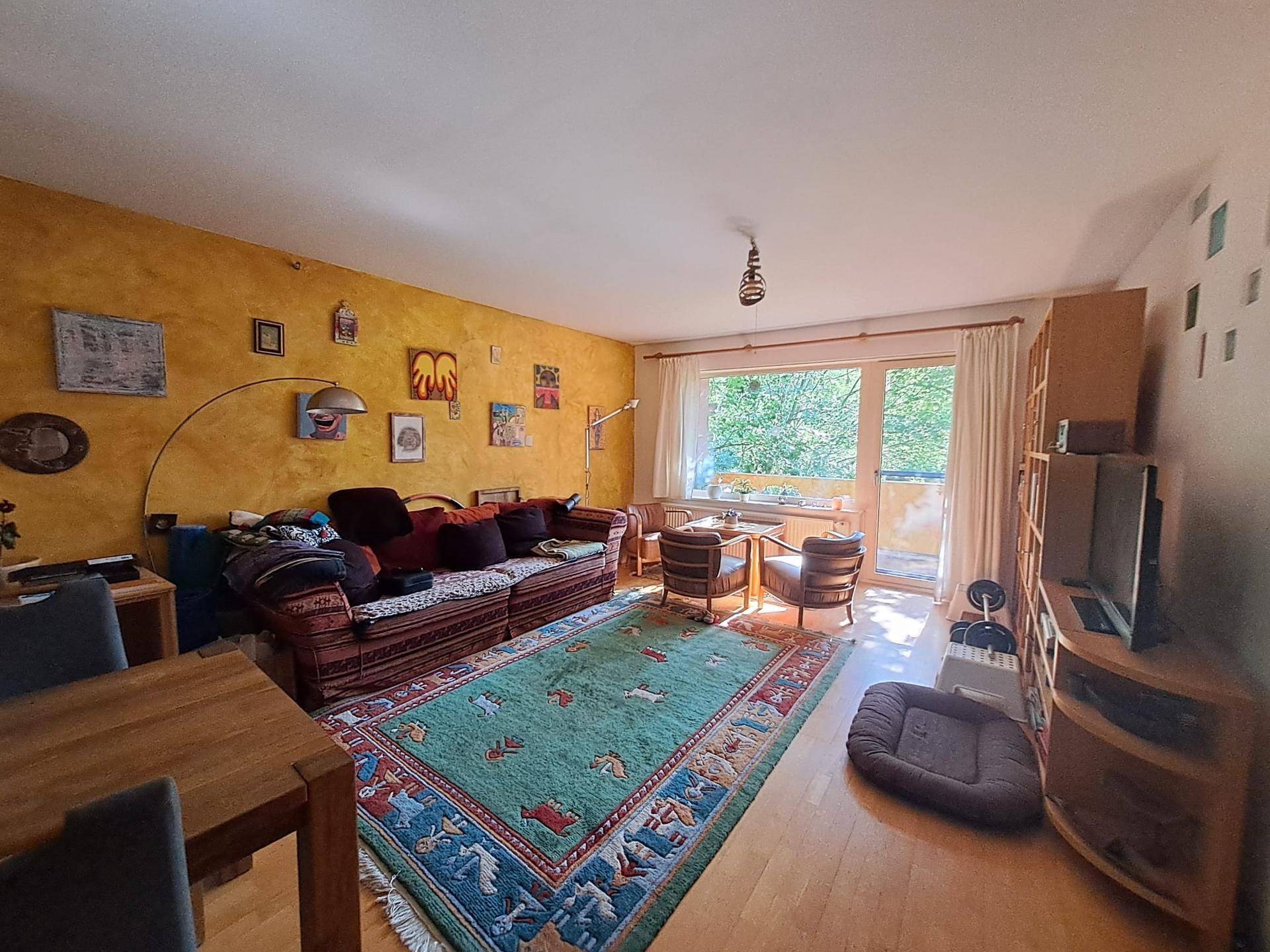 Geräumiger Wohnbereich mit gemütlichem Sofa, farbenfrohem Teppich und Zugang zum Balkon mit Grünblick.