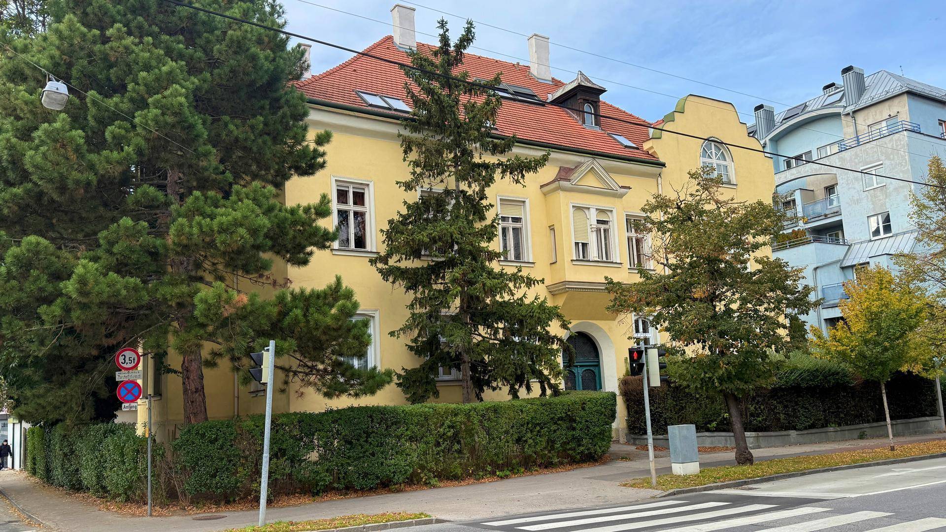 Gelbes Mehrfamilienhaus mit rotem Ziegeldach, umgeben von Bäumen und Hecken an einer Straße.