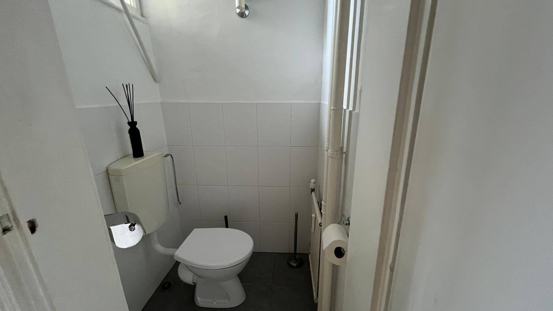 Separates WC mit Toilette, weiß gefliesten Wänden und dunklem Boden in schlichtem Stil.
