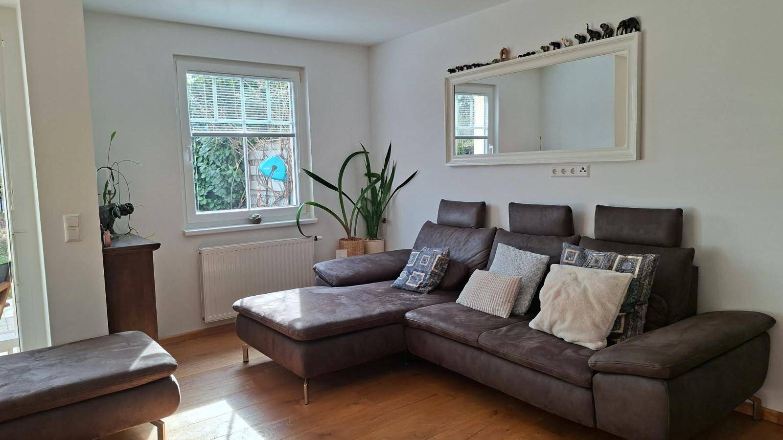 Gemütliches Wohnzimmer mit großem Ecksofa, Holzboden und Fenster mit Blick ins Grüne.