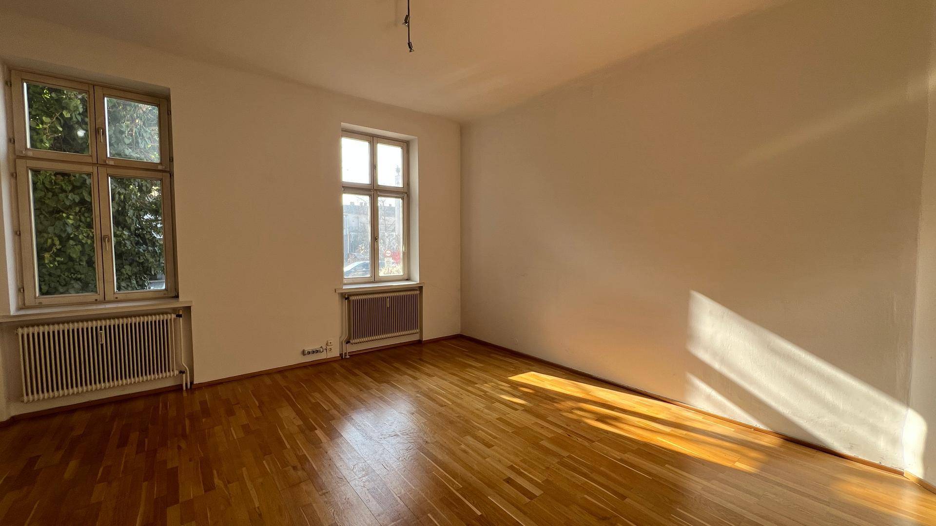 Geräumiges, helles Zimmer mit Holzparkettboden und großen Fenstern, die viel Sonnenlicht hereinlassen.