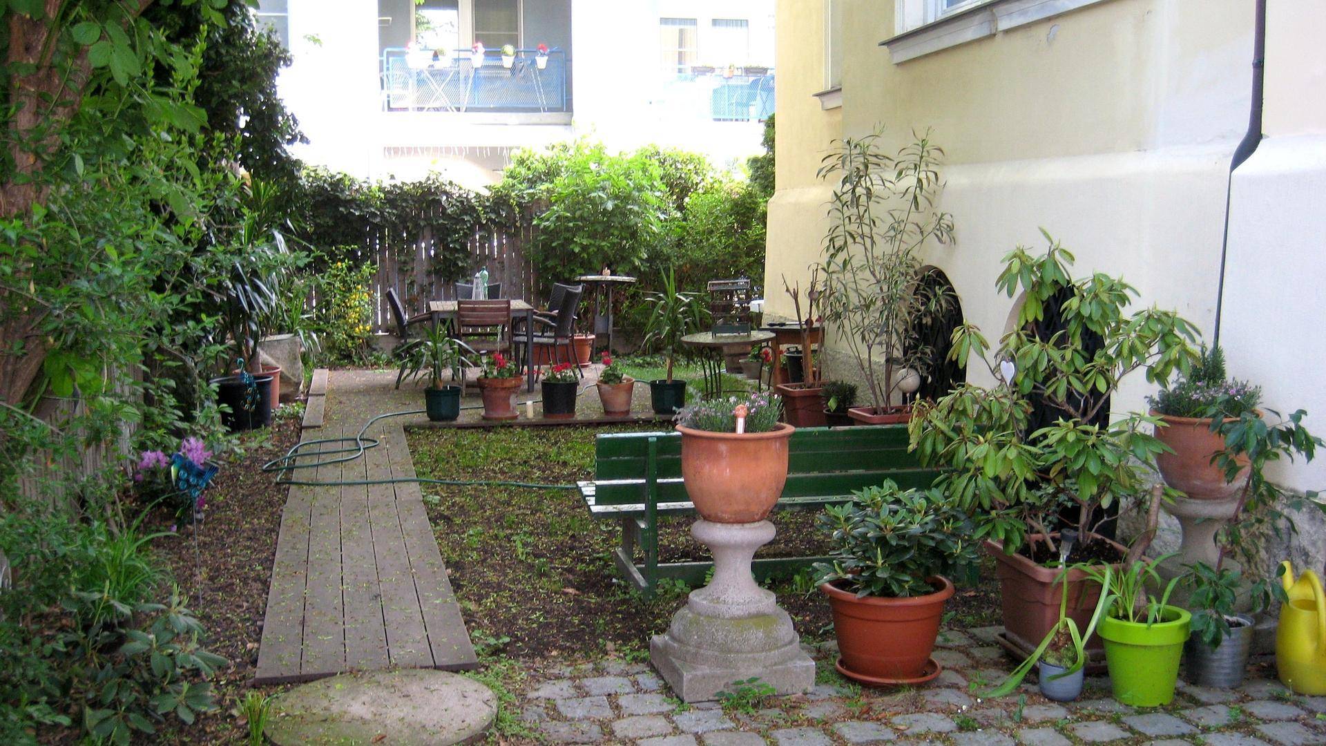 Grüner Hinterhofgarten mit Sitzbereich, vielen Topfpflanzen und einem Holzbodenweg.
