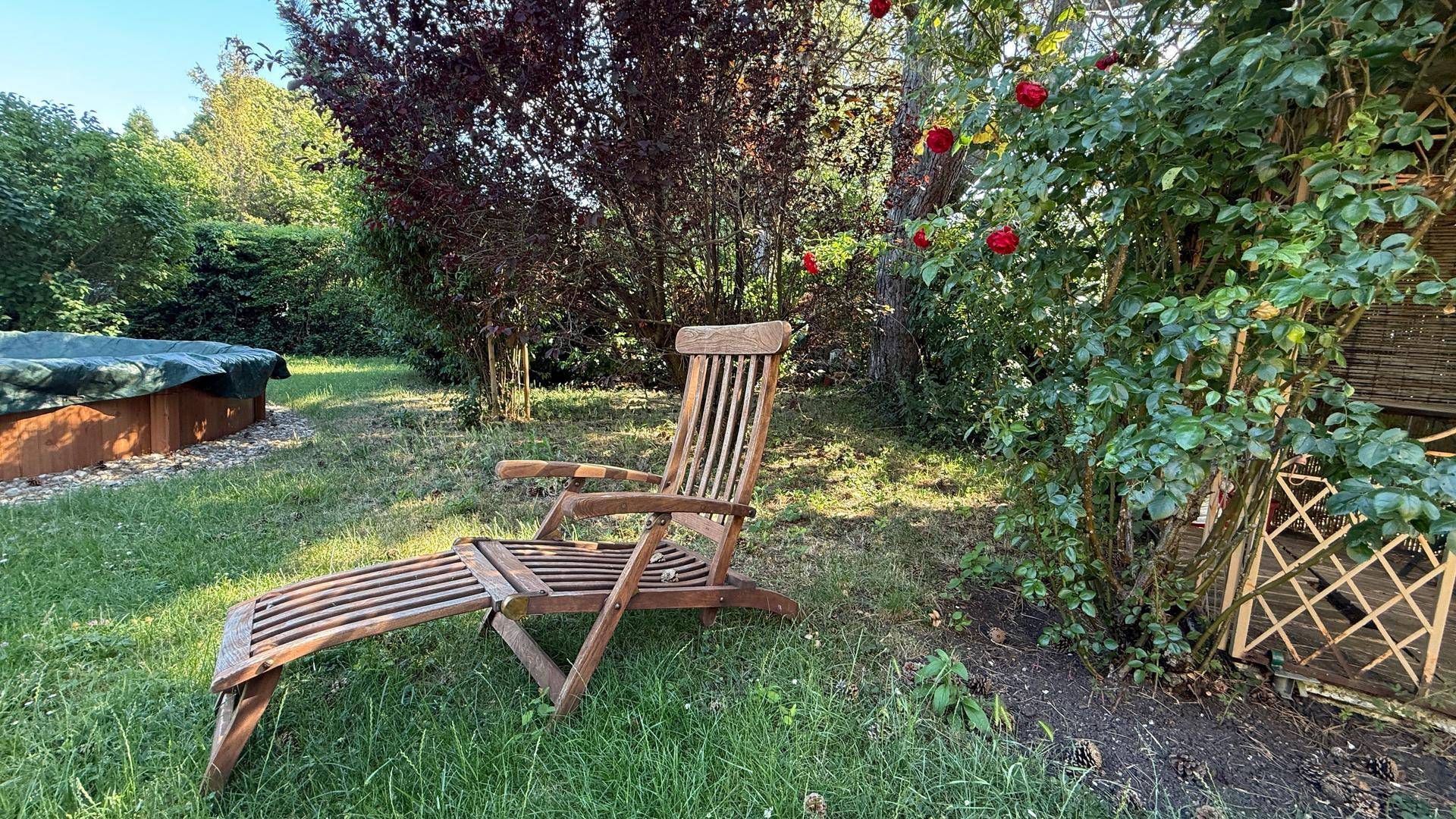 Entspannungsbereich im Garten mit Holzliege und einem abgedeckten Whirlpool unter Bäumen.