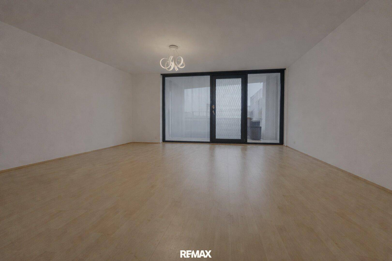 Geräumiges, leeres Wohnzimmer mit hellem Laminatboden und Zugang zum Balkon.