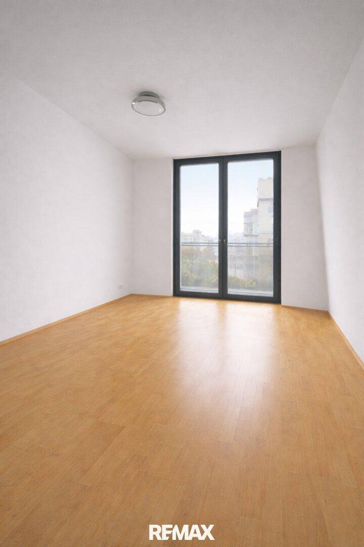 Leeres Zimmer mit Laminatboden und großem Fenster, das viel Licht hereinlässt.