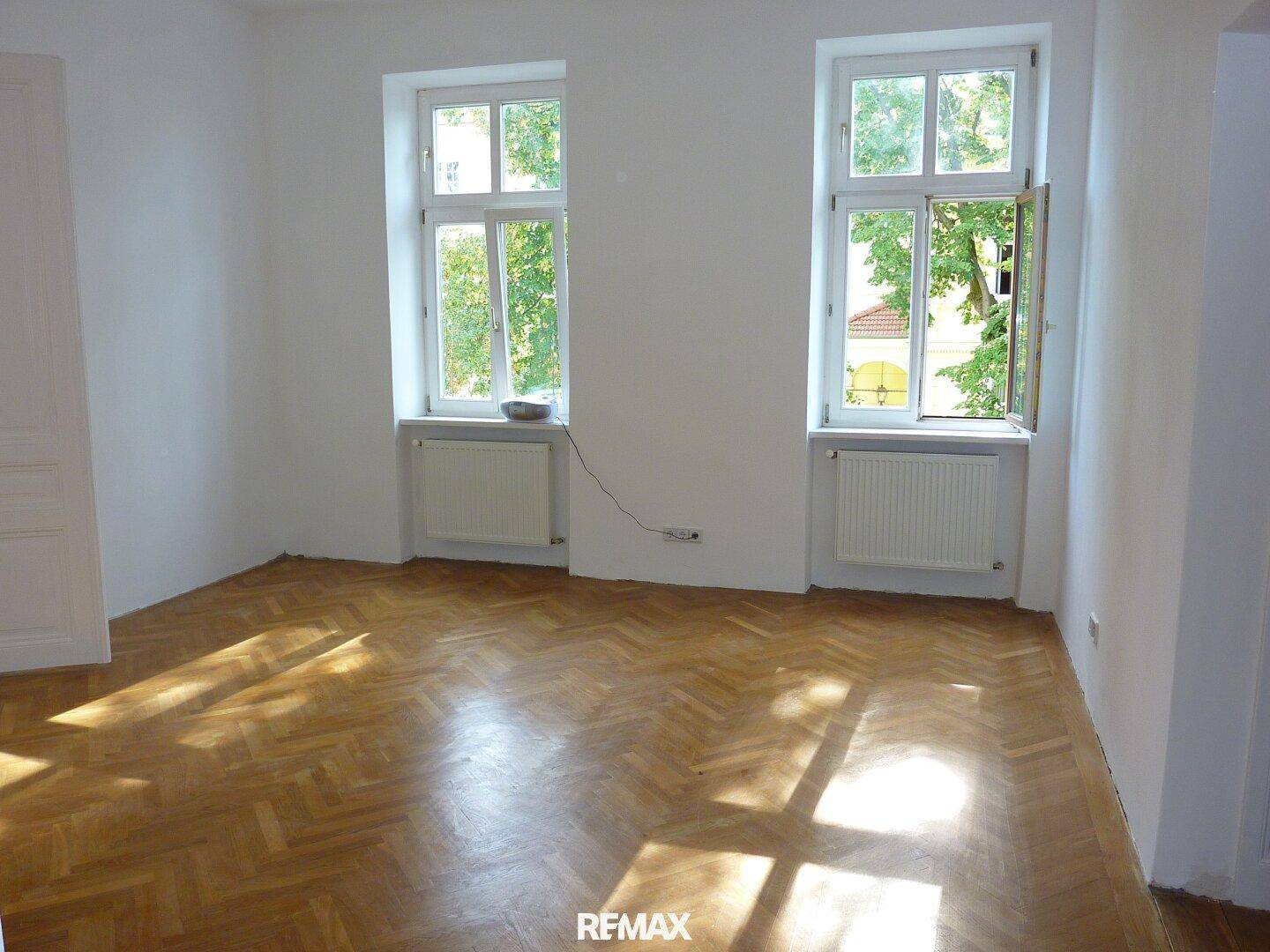 Lichtdurchflutetes Zimmer mit klassischem Fischgrätparkett und Blick ins Grüne durch geöffnete Fenster.