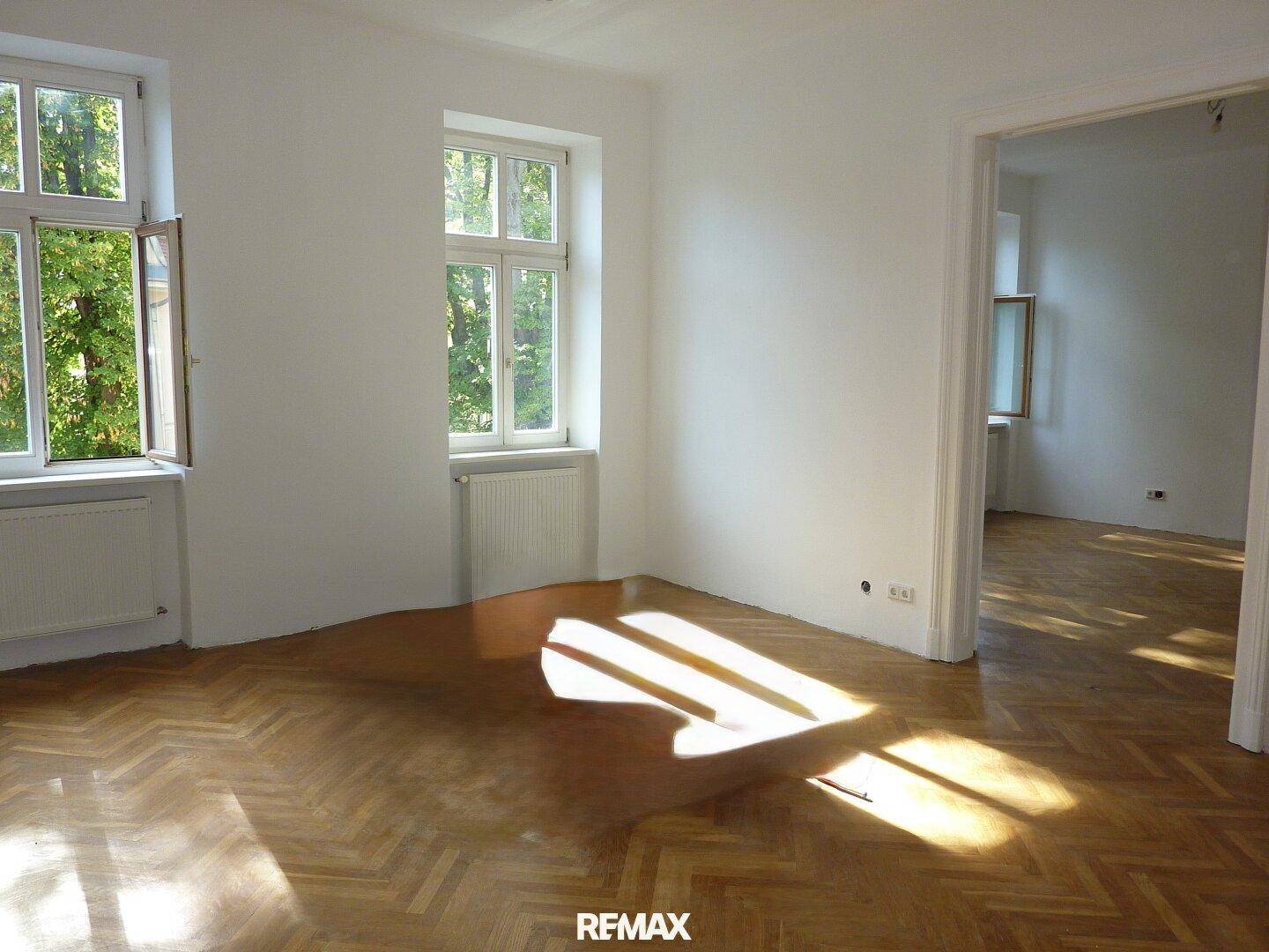 Helles Zimmer mit Fischgrätparkettboden, weißen Wänden und viel natürlichem Licht durch große Fenster.