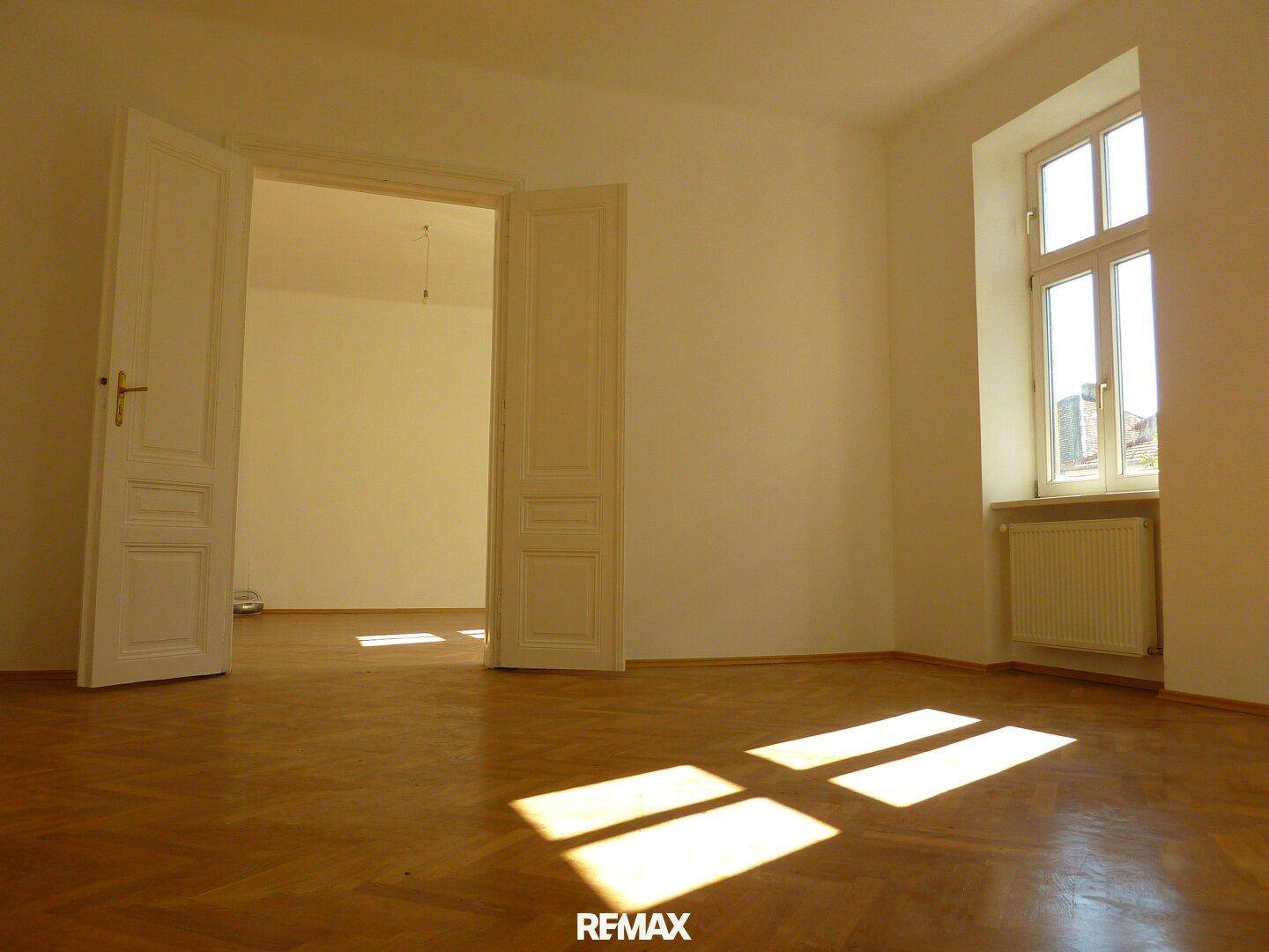 Geräumiges Zimmer mit klassischem Fischgrätparkett und doppelten Flügeltüren zu einem weiteren Raum.