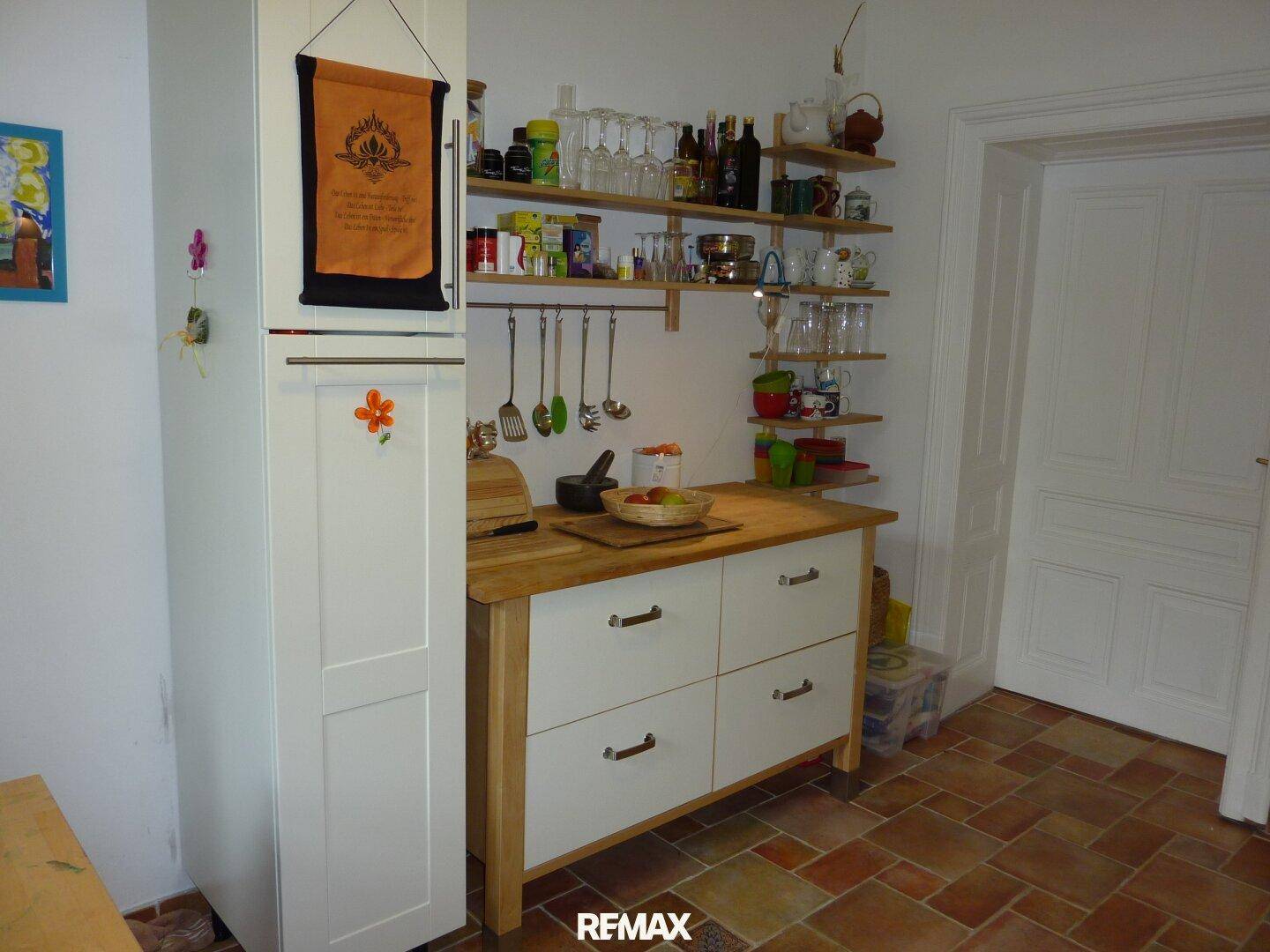 Küchenbereich mit Kühlschrank, Holz-Arbeitsplatte, offenen Regalen und Terrakotta-Fliesenboden.