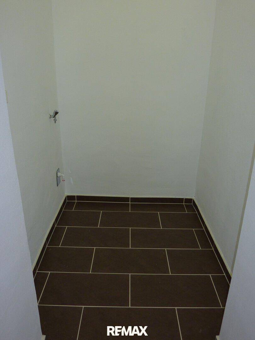 Kleiner Abstellraum mit dunklen Fliesen und Anschlüssen, ideal für Waschmaschine oder Lagerung.
