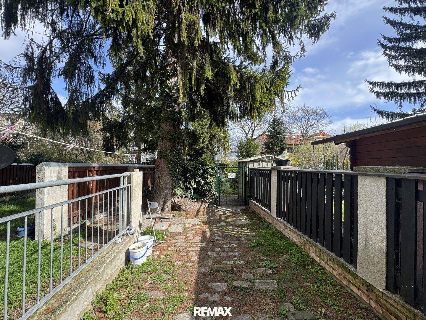 Gartenweg mit altem Baum, umzäuntem Bereich und einem kleinen Gartenhaus im Hintergrund.