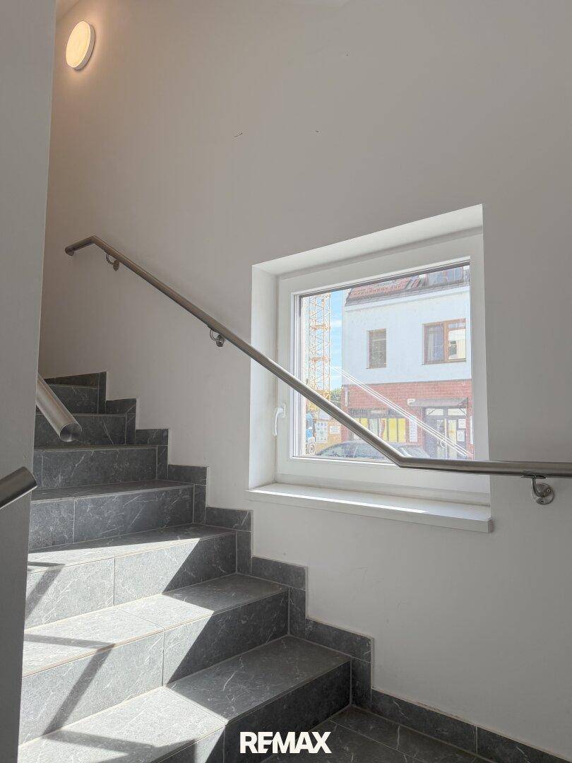 Treppenhaus mit Fliesenstufen, Edelstahlgeländer und Fenster für natürliches Licht.