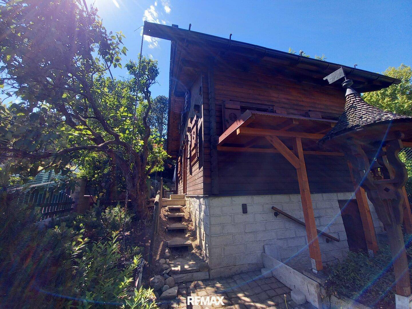Charmantes Holzhaus mit Steinfundament und Treppenzugang im Grünen unter strahlendem Sonnenschein.