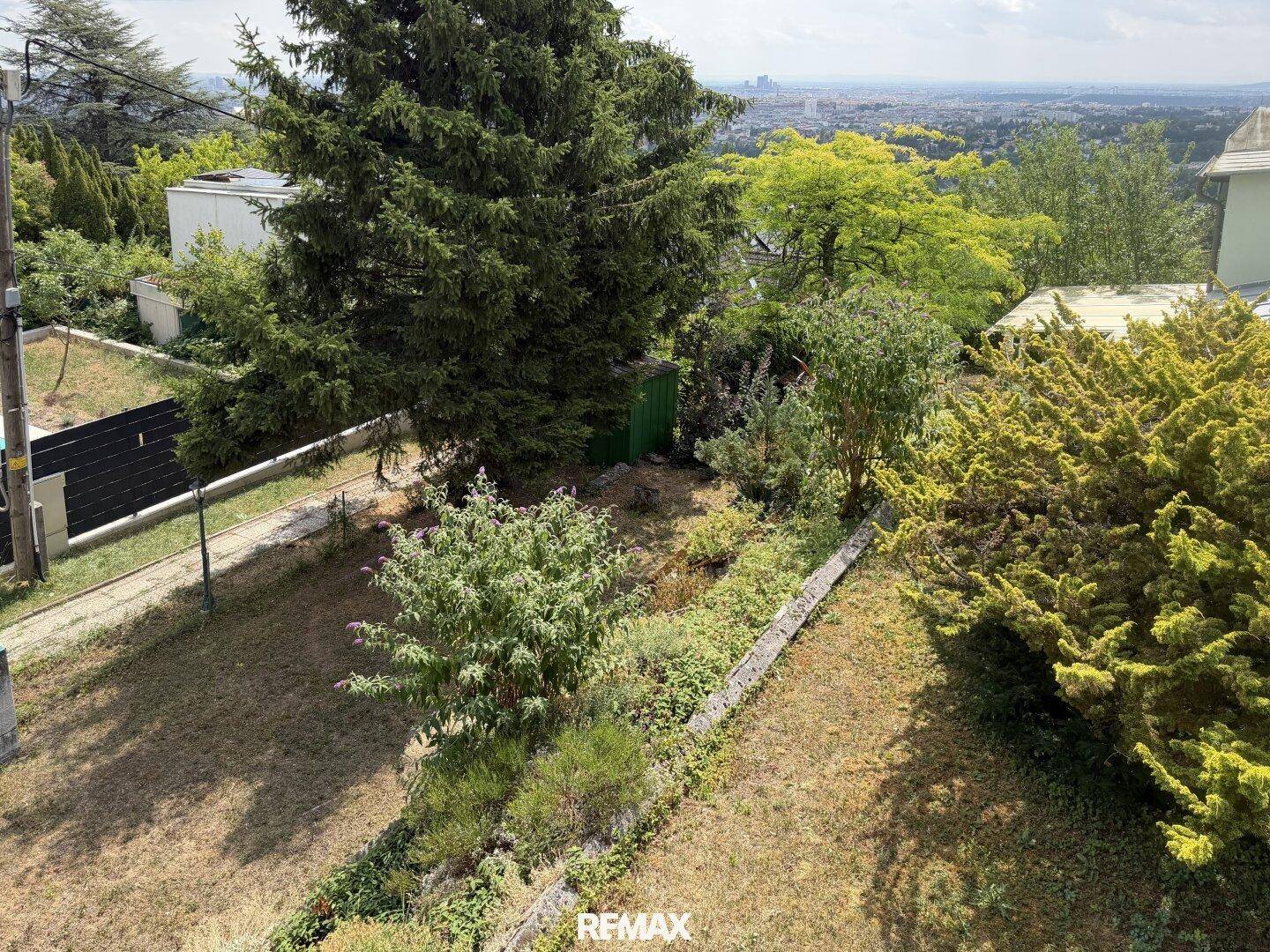 Gartenbereich mit verschiedenen Pflanzen, Bäumen und einem Blick auf die Stadt in der Ferne.
