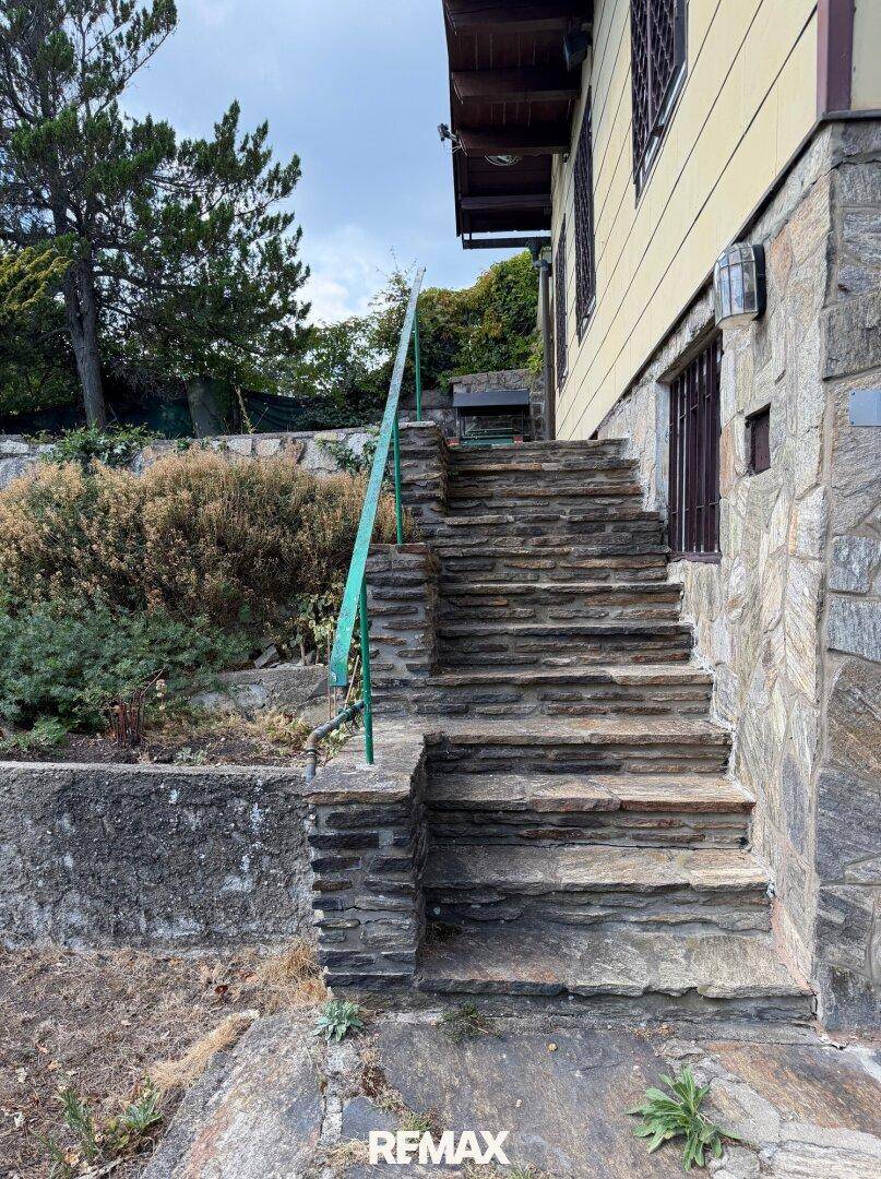 Steintreppe mit grünem Geländer, die zum Haus führt, umgeben von natürlicher Vegetation.