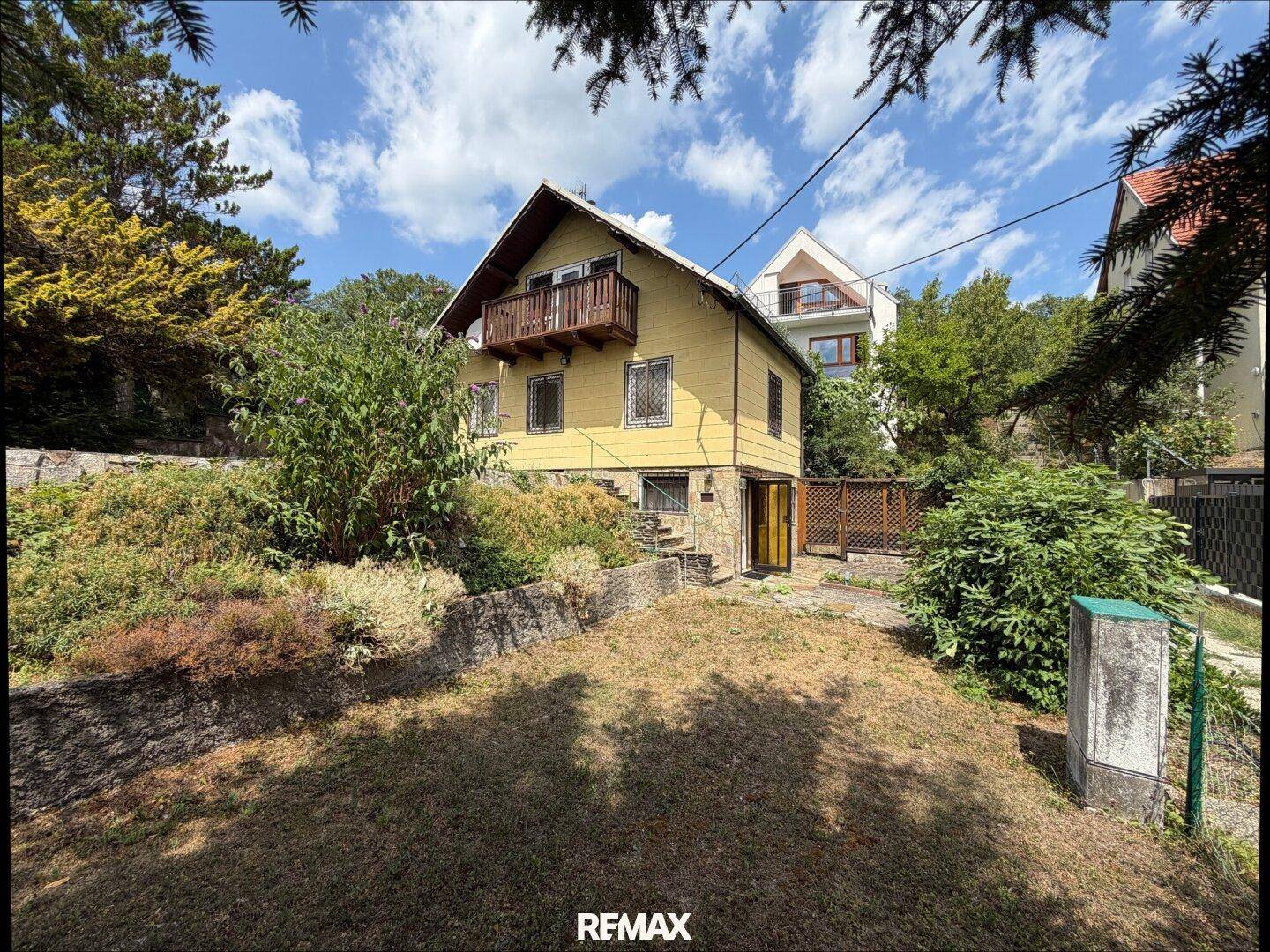 Freistehendes Haus mit Balkon und Garten in Hanglage, umgeben von Bäumen und Sträuchern.