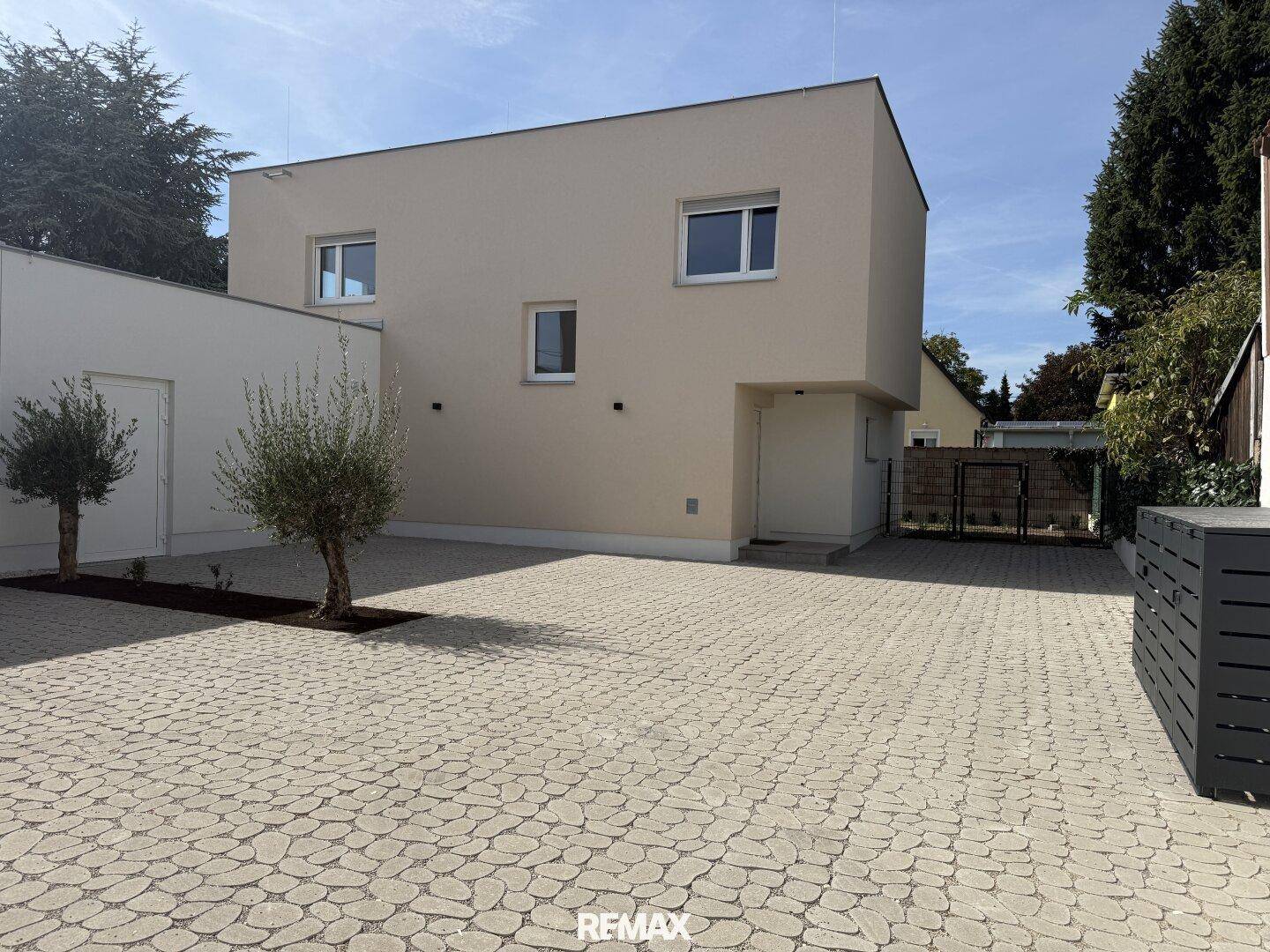 Moderne Neubaufassade mit beigefarbenem Putz, großen Fenstern und gepflasterter Auffahrt.