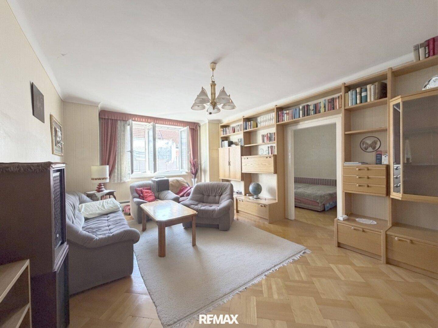 Helles Wohnzimmer mit Parkettboden und Blick in ein angrenzendes Schlafzimmer, ausgestattet mit Bücherregalen.