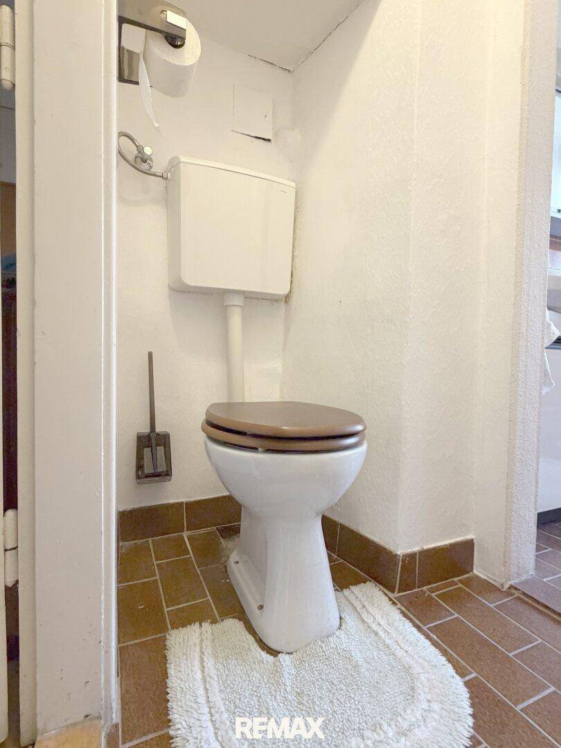 Separates WC mit einfacher Toilette und braunen Bodenfliesen, funktional und schlicht gehalten.