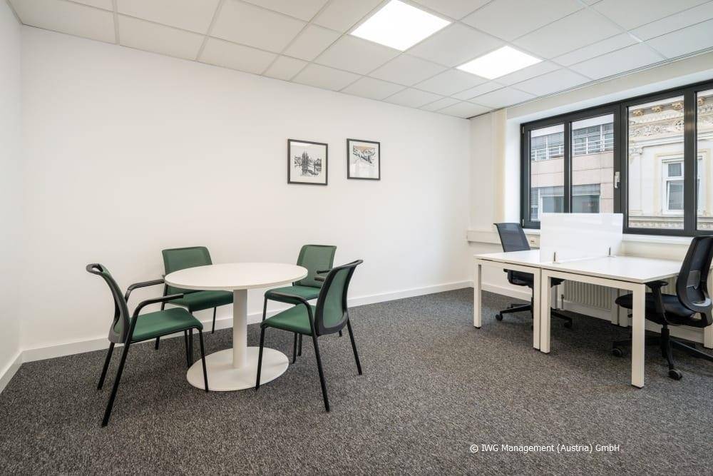 Helles Büro mit Teppichboden, einem kleinen Besprechungstisch und zwei Arbeitsplätzen am Fenster.