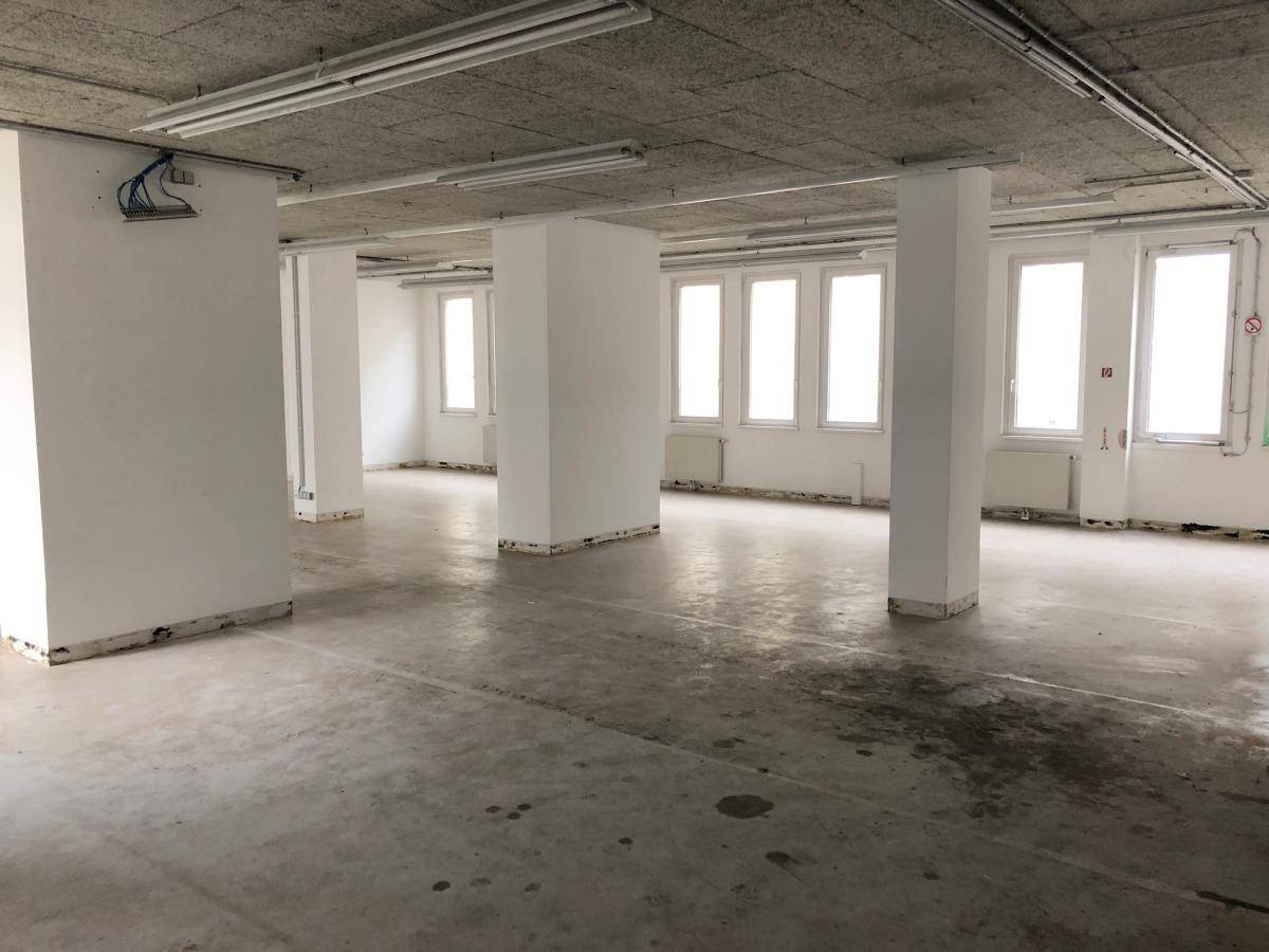 Geräumiger, unmöblierter Bürobereich mit Säulen, vielen Fenstern und sichtbaren Installationen an der Decke.