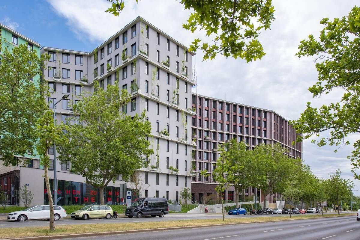 Straßenansicht moderner Gebäude mit unterschiedlichen Fassaden, parkenden Autos und einer Reihe von Bäumen entlang der Straße.