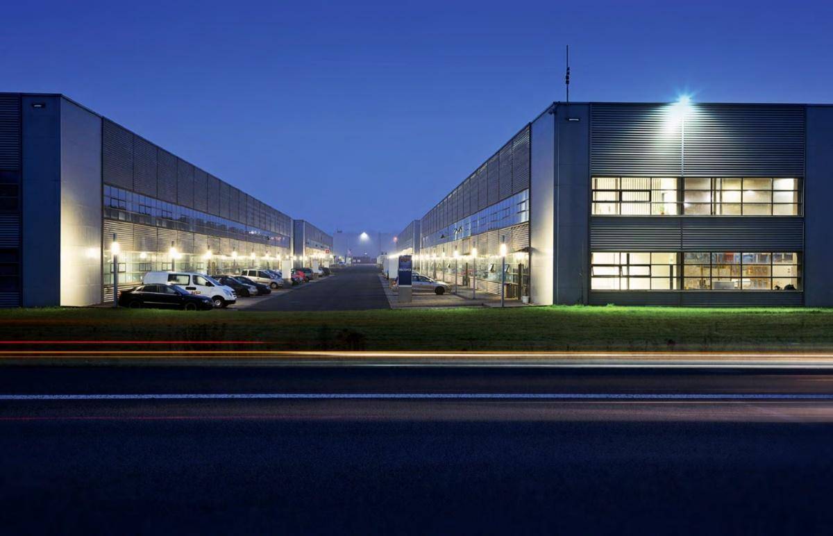 Industriegebäude bei Dämmerung mit beleuchteten Fassaden und Parkplätzen entlang einer Straße.