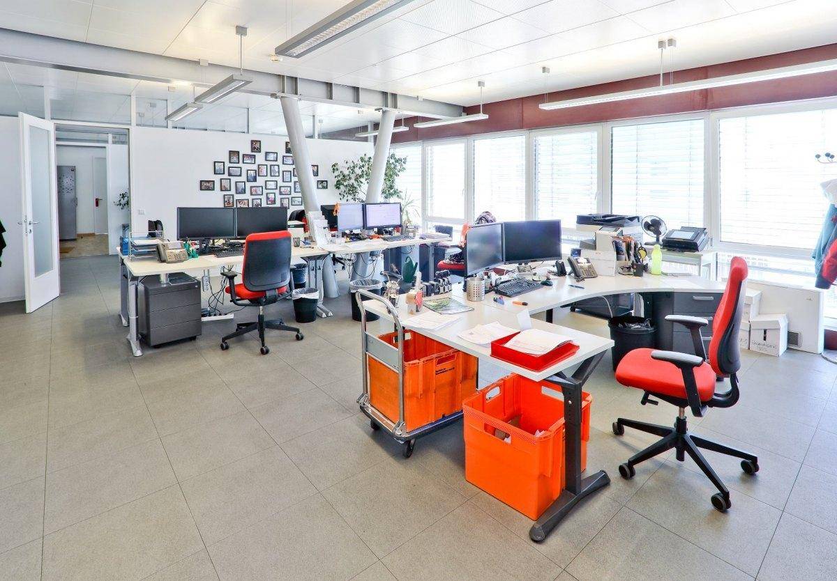 Weitläufiges Büro mit mehreren Arbeitsplätzen, roten Bürostühlen und viel Tageslicht durch große Fenster.