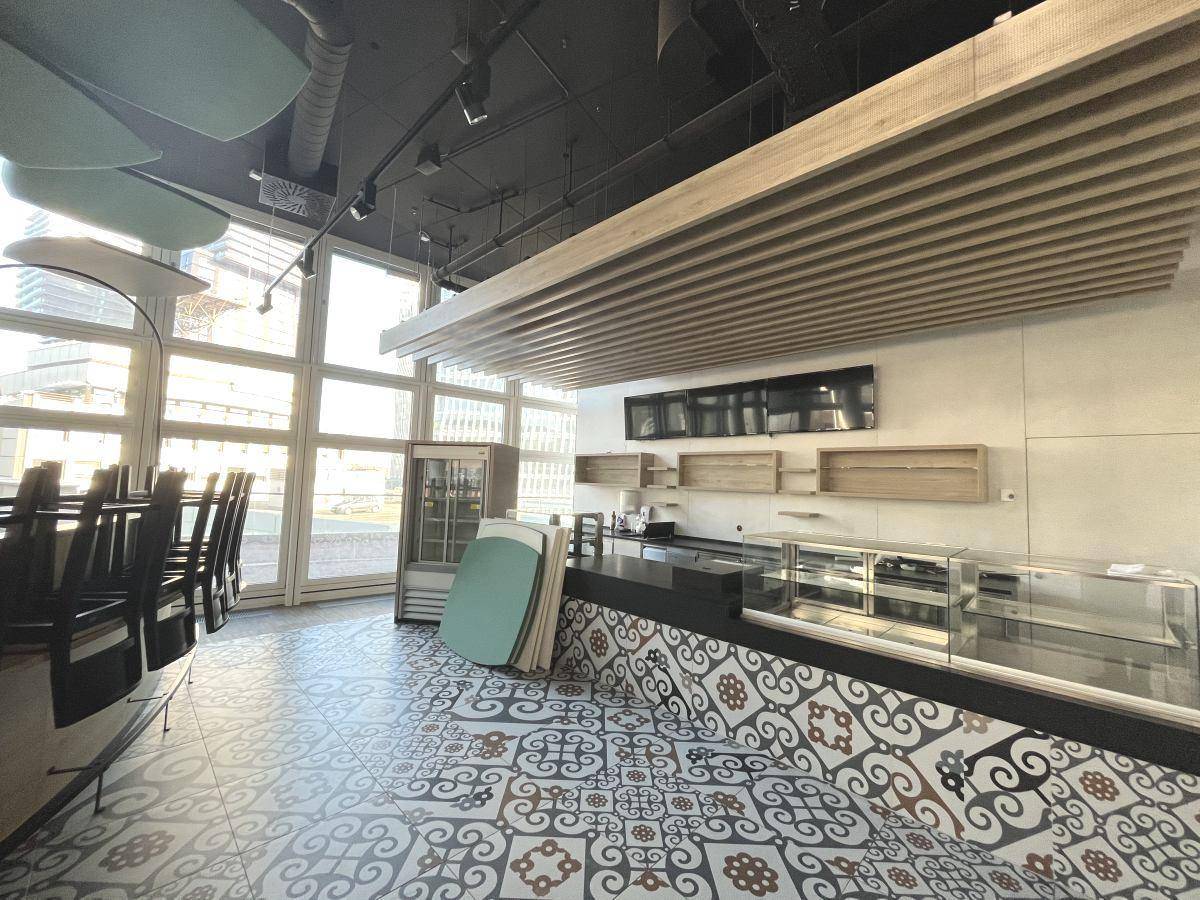 Thekenbereich eines Cafés mit Auslagen, dunklen Möbeln und großen Fenstern.
