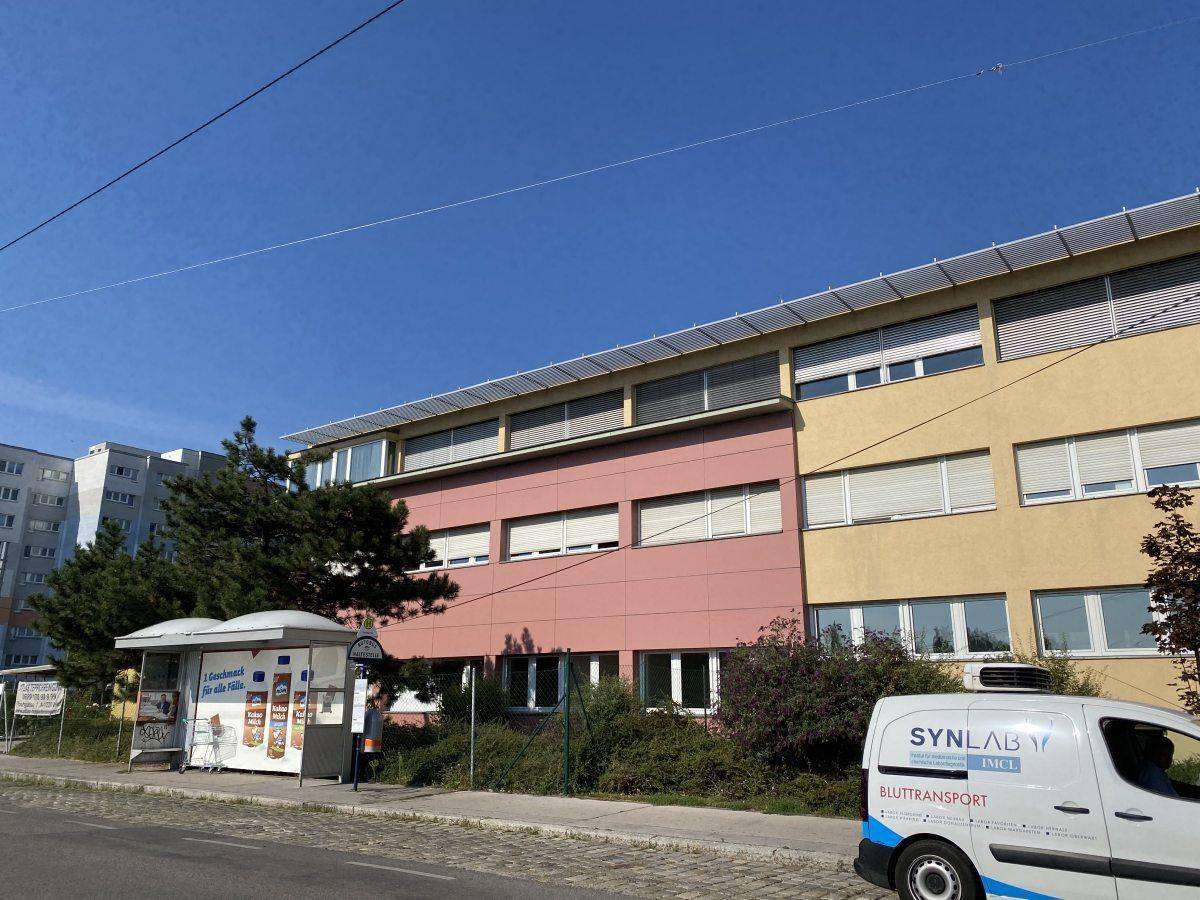 Gebäudeansicht mit gelber und rosafarbener Fassade, umgeben von Büschen und einem Buswartehäuschen.