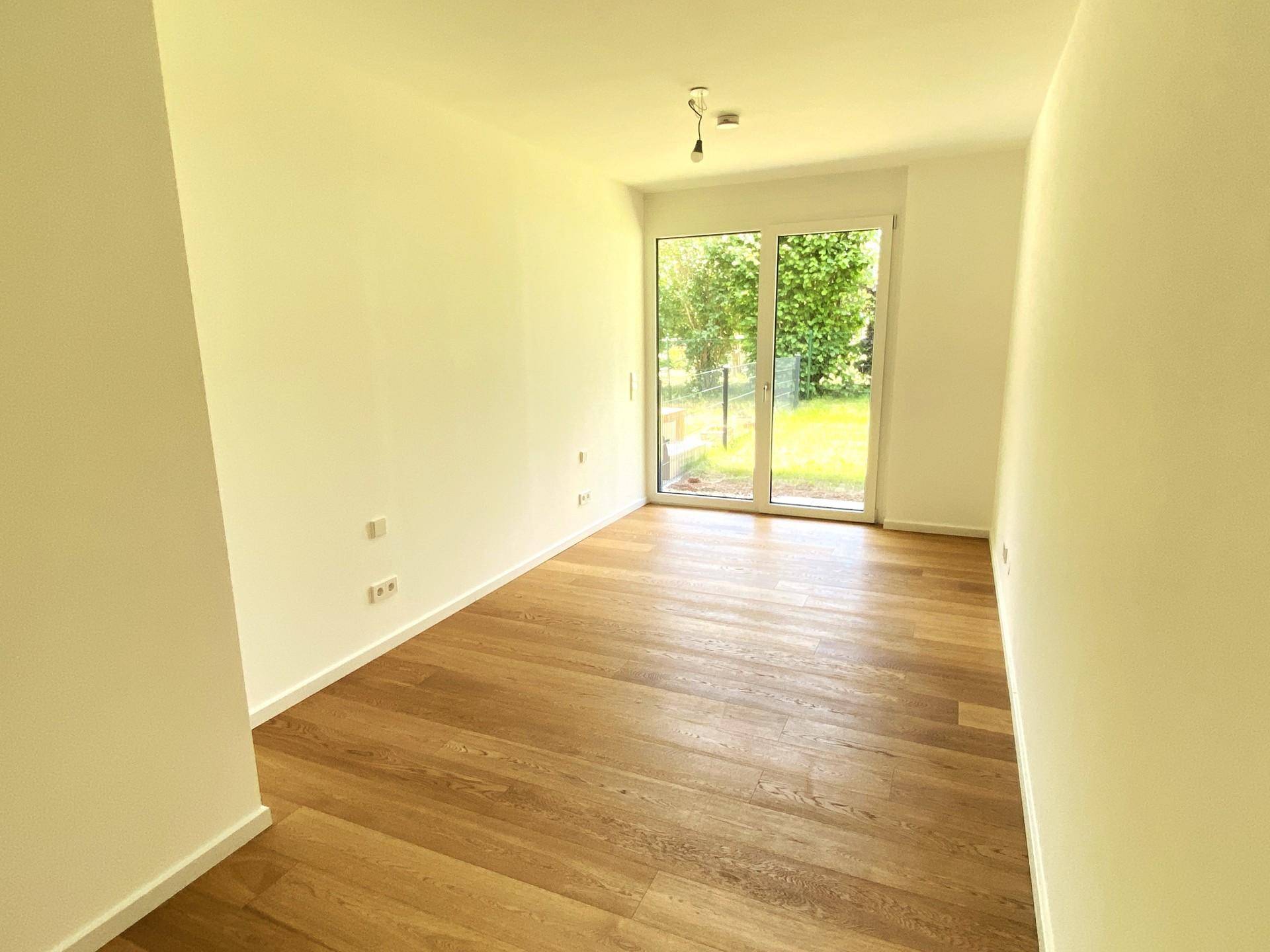 Heller, leerer Raum mit Holzboden und großem Fenster, das Zugang zu einem grünen Garten bietet.