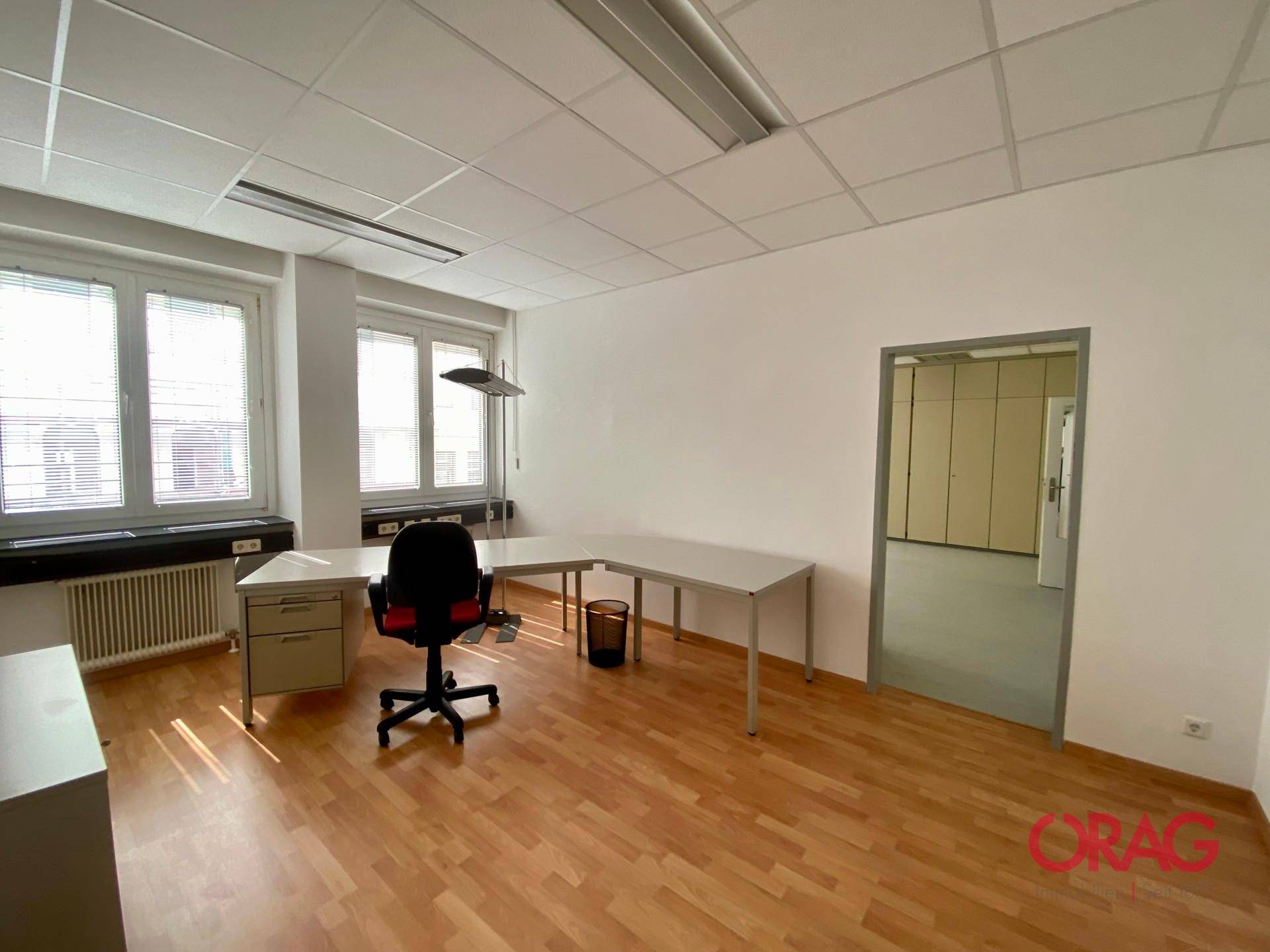 Geräumiges Büro mit Holzparkettboden, Schreibtisch und Blick in einen weiteren Raum.