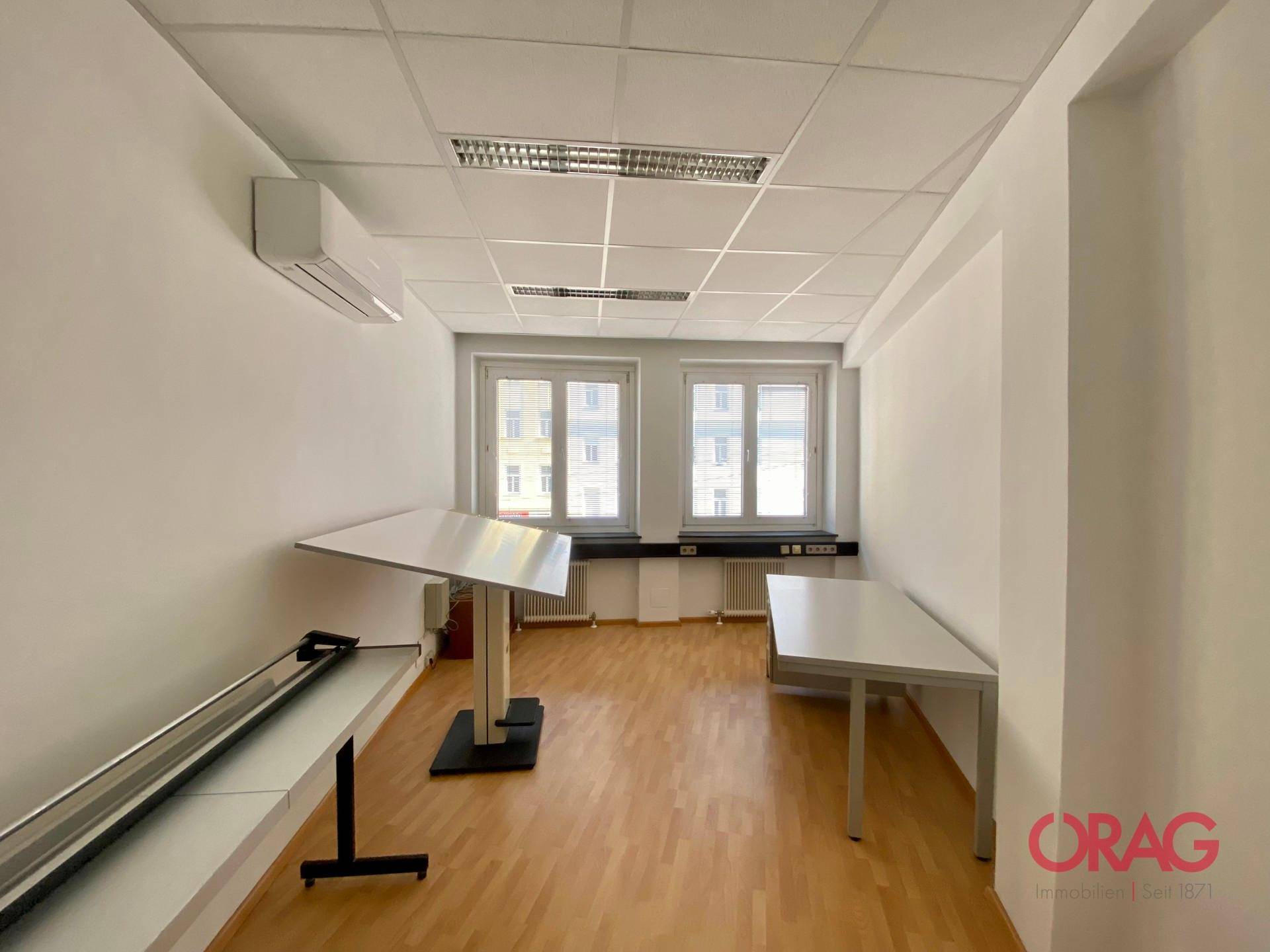 Helles Büro mit Holzparkettboden, zwei Fenstern und Klimaanlage an der Wand.