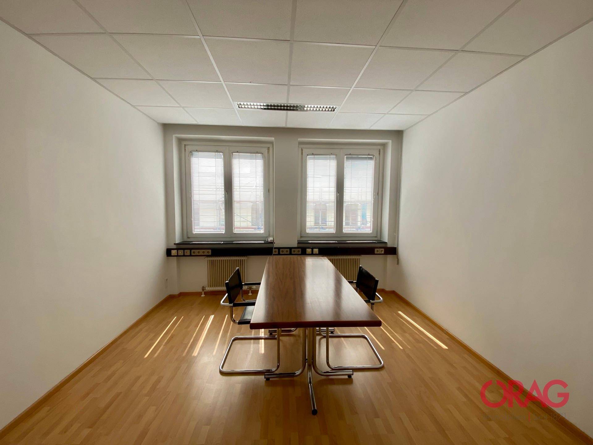 Schlichter Büroraum mit Holzparkettboden, zwei Fenstern und einem langen Tisch.