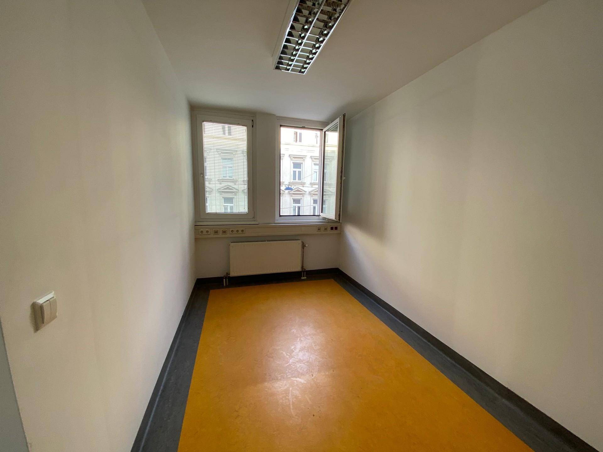 Kleinerer Raum mit auffälligem gelbem Boden, zwei Fenstern und einem Heizkörper, ideal für ein Einzelbüro.