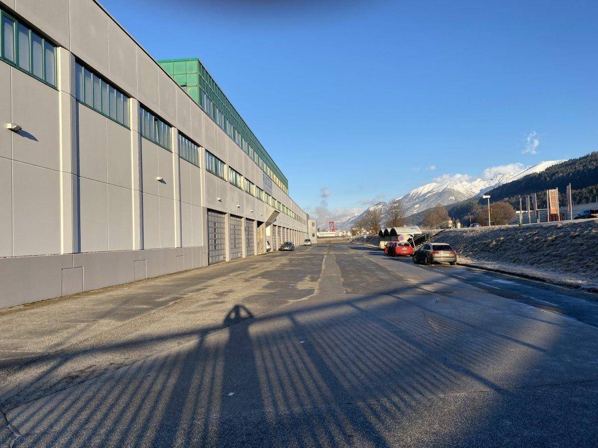 Seitenansicht der Industriehalle mit breiter Zufahrt und geparkten Fahrzeugen, im Hintergrund schneebedeckte Berge.