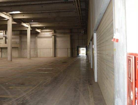 Lange, leere Lagerhalle mit Betonstützen und einem Gang, der zu einem Ausgang führt.