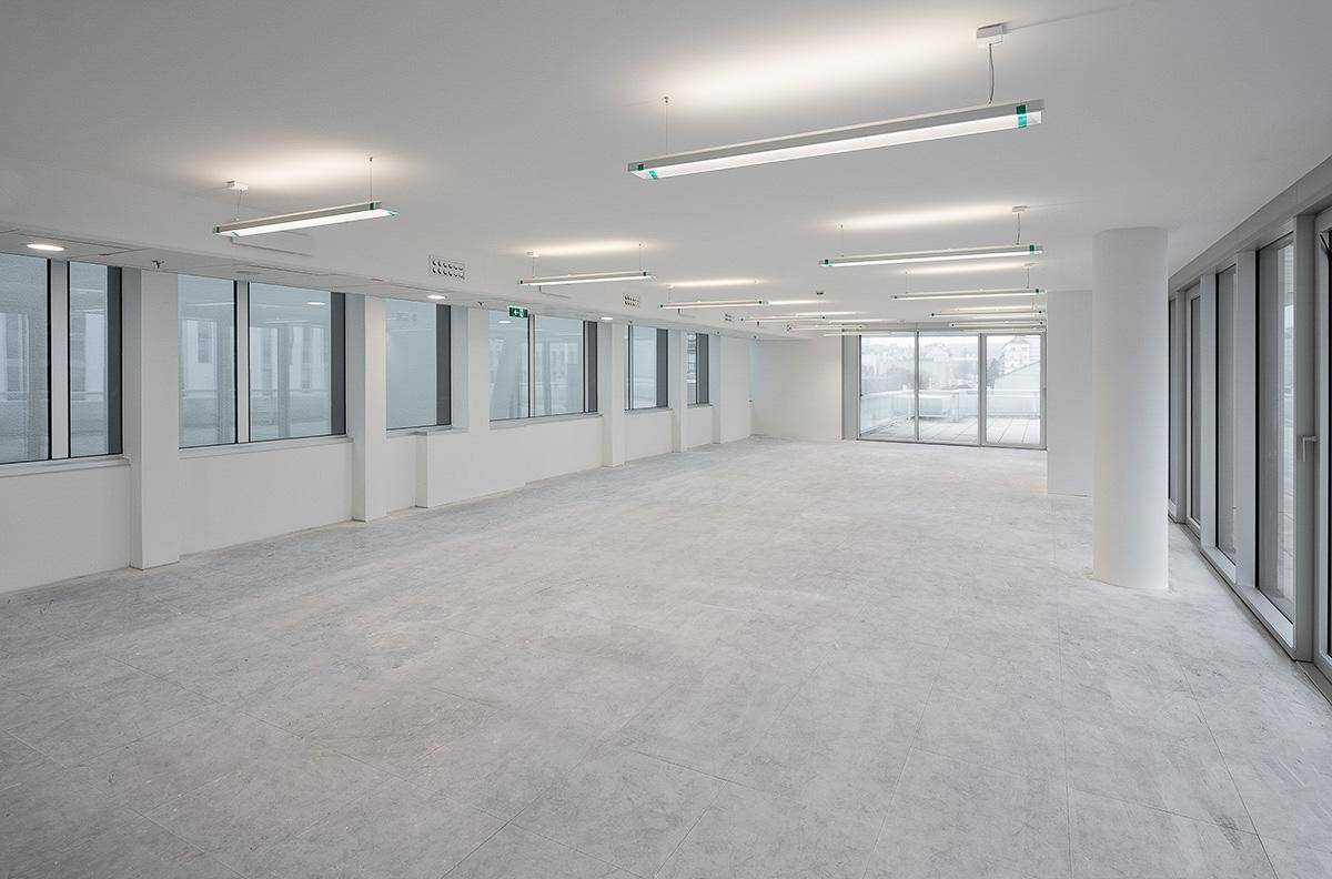 Weitläufiges, leeres Büro mit hellem Betonboden und großen Fenstern, bietet viel natürliches Licht.