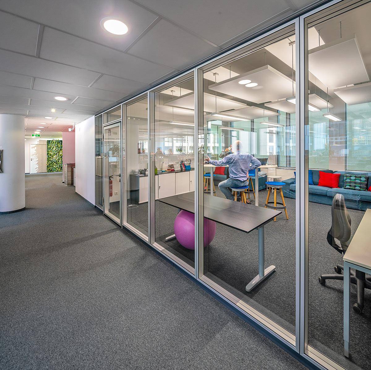 Moderner Bürobereich mit Glaswänden, Teppichboden und farbenfrohen Möbeln, ideal für kreatives Arbeiten.