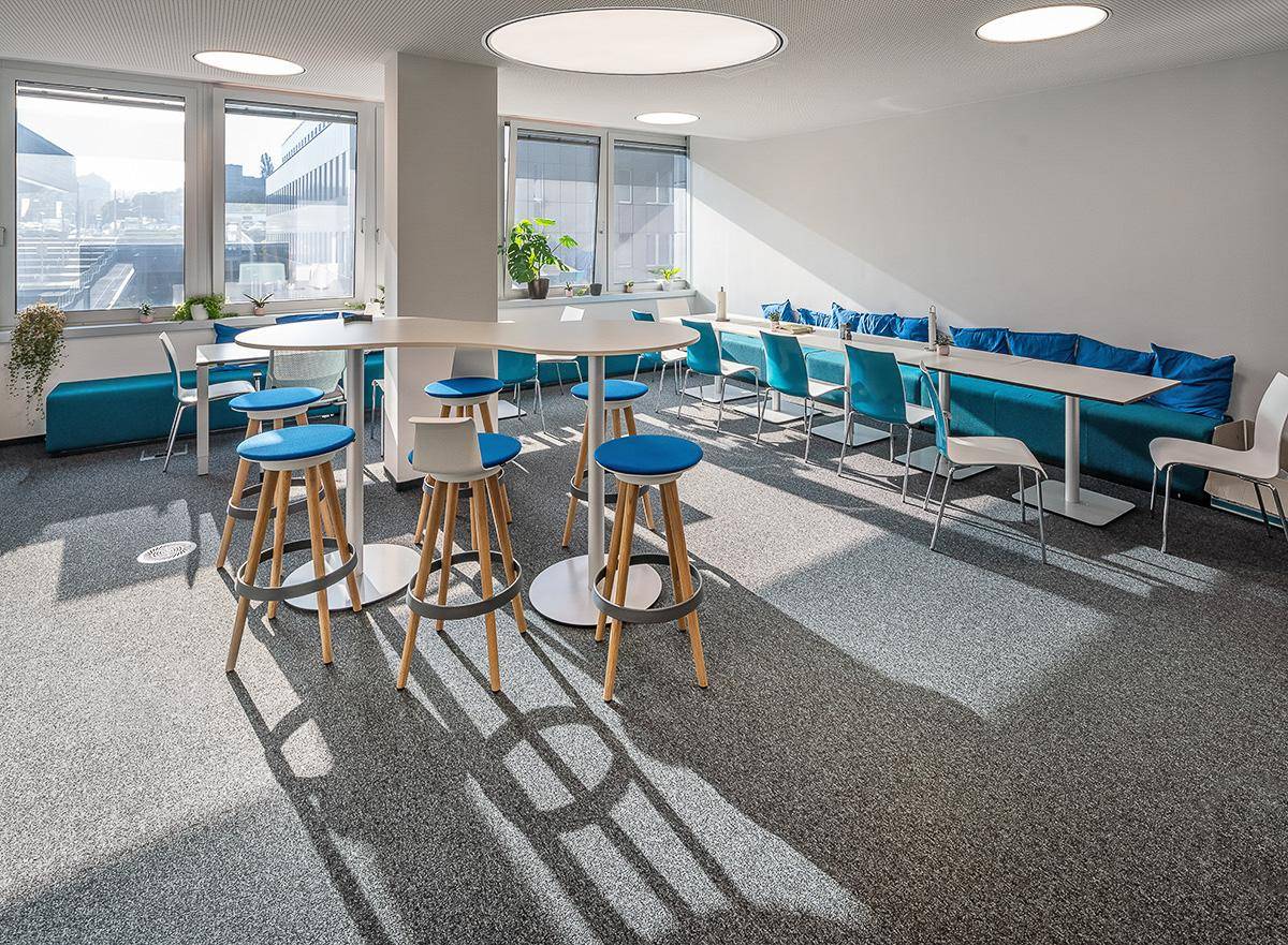 Heller Pausenbereich im Büro mit hohen Tischen, Hockern und blauen Sitzgelegenheiten, ideal für informelle Treffen.