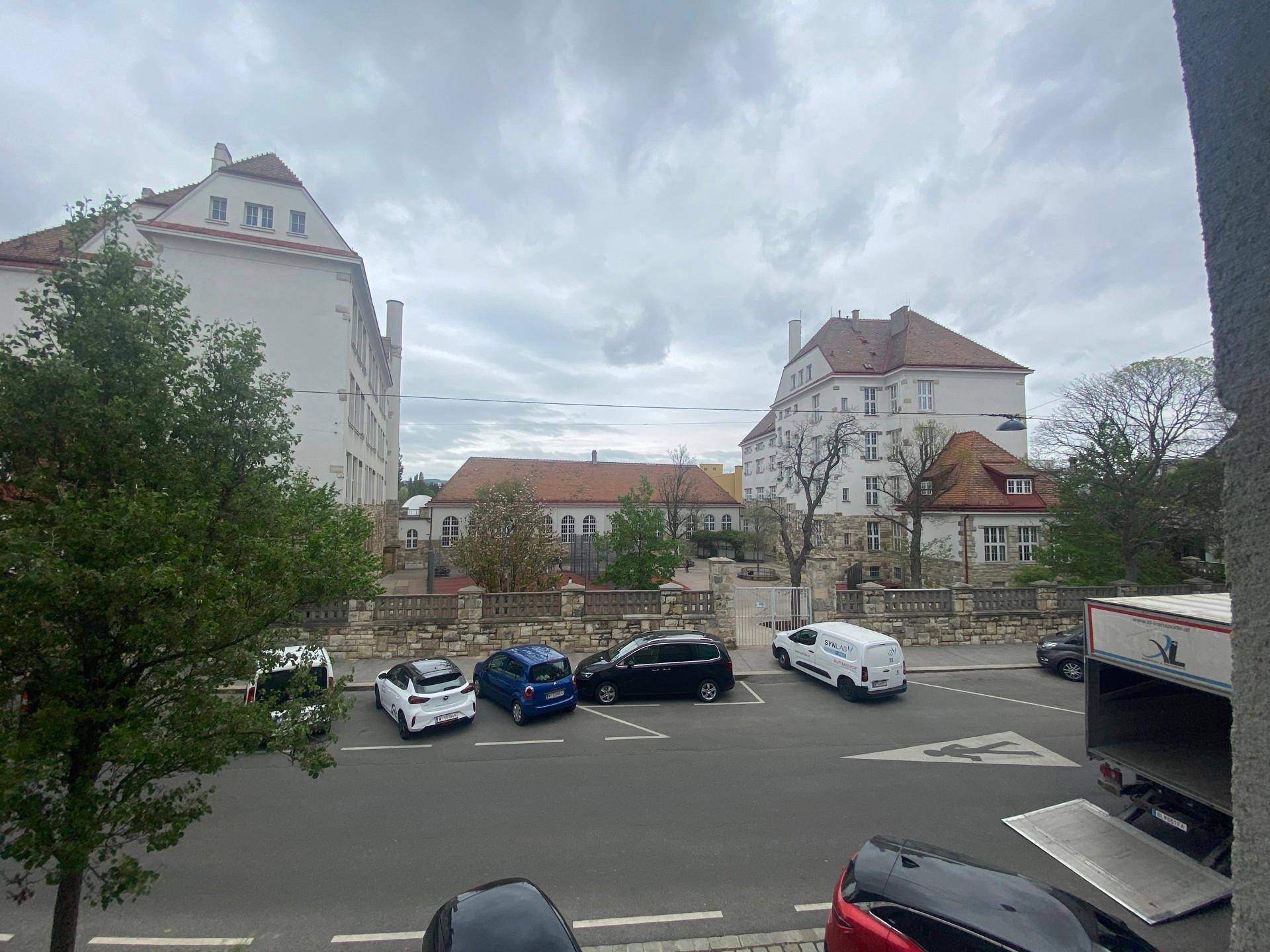 Blick aus dem Fenster auf umliegende Gebäude, eine Straße mit geparkten Autos und grüne Bäume.